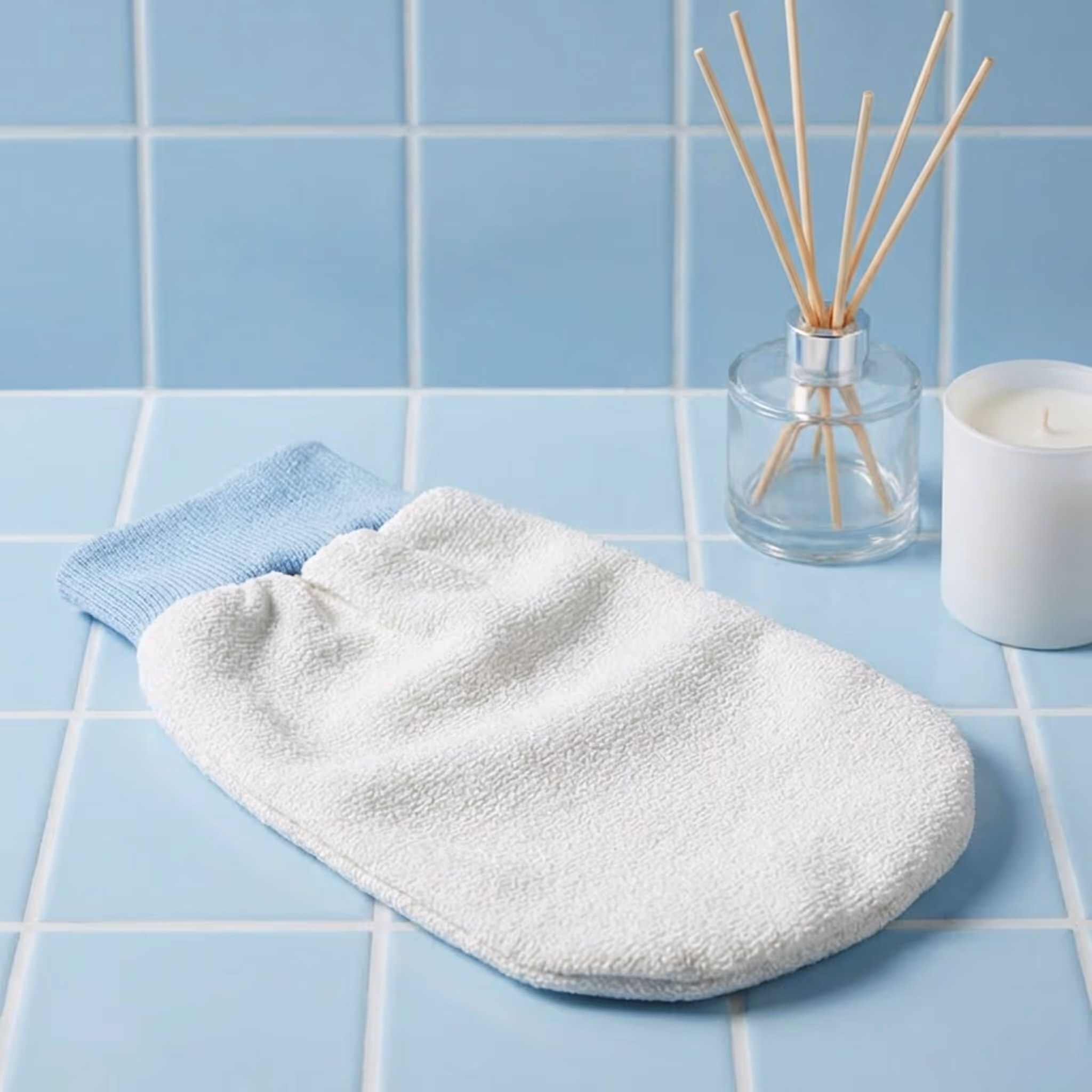 White After Spa Exfoliating Body Mitt placed on blue tiled surface beside reed diffuser and candle.