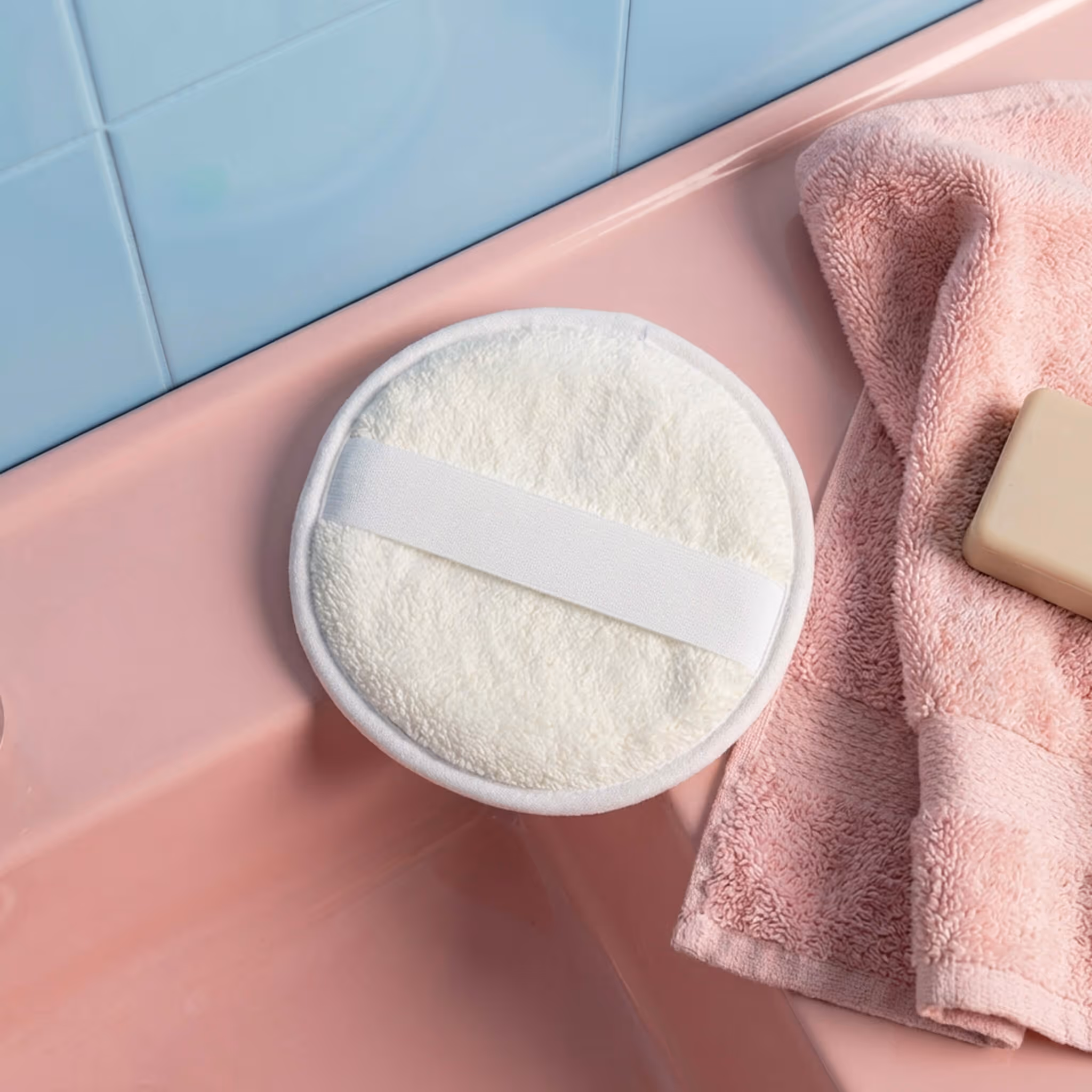 After Spa Dual Texture Scrubber resting on pink bathtub edge beside towel and soap.