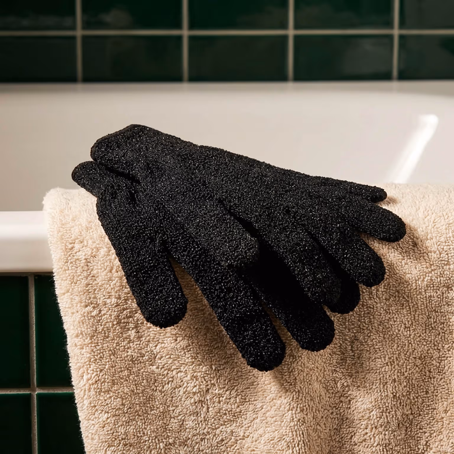 Black exfoliating shower gloves resting on beige towel near bathtub in bathroom interior