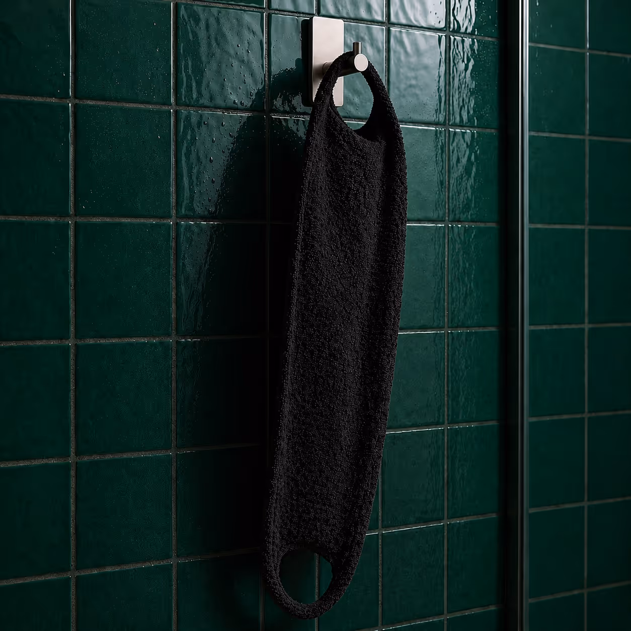 Black exfoliating back scrubber hanging on wall hook in shower with dark green tile background