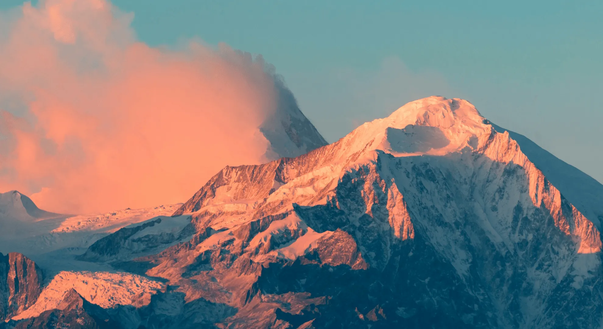 Snow-covered mountain peaks lit by warm orange sunlight with pink clouds and clear blue sky.