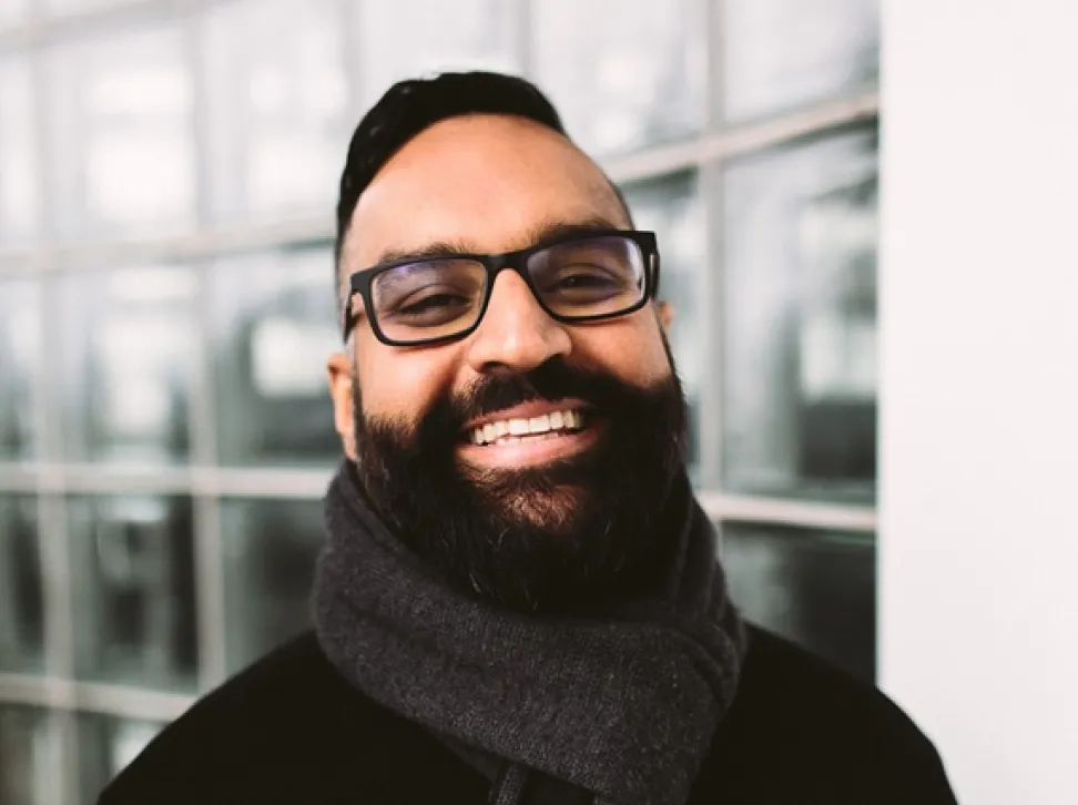 Smiling Gordy Bal with beard, glasses, and a dark scarf standing in front of a windowed wall.