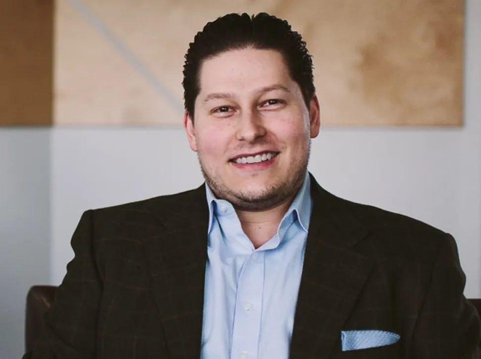 Smiling Aaron Samsonoff with dark slicked-back hair wearing a dark blazer over a light blue shirt.