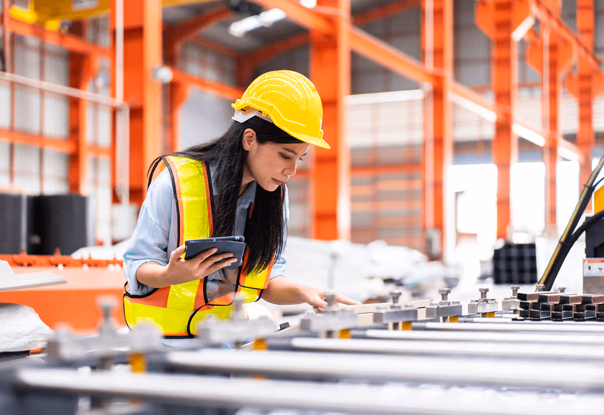 Woman in a yellow hard hat and safety vest inspecting metal bars in an industrial warehouse while holding a tablet.