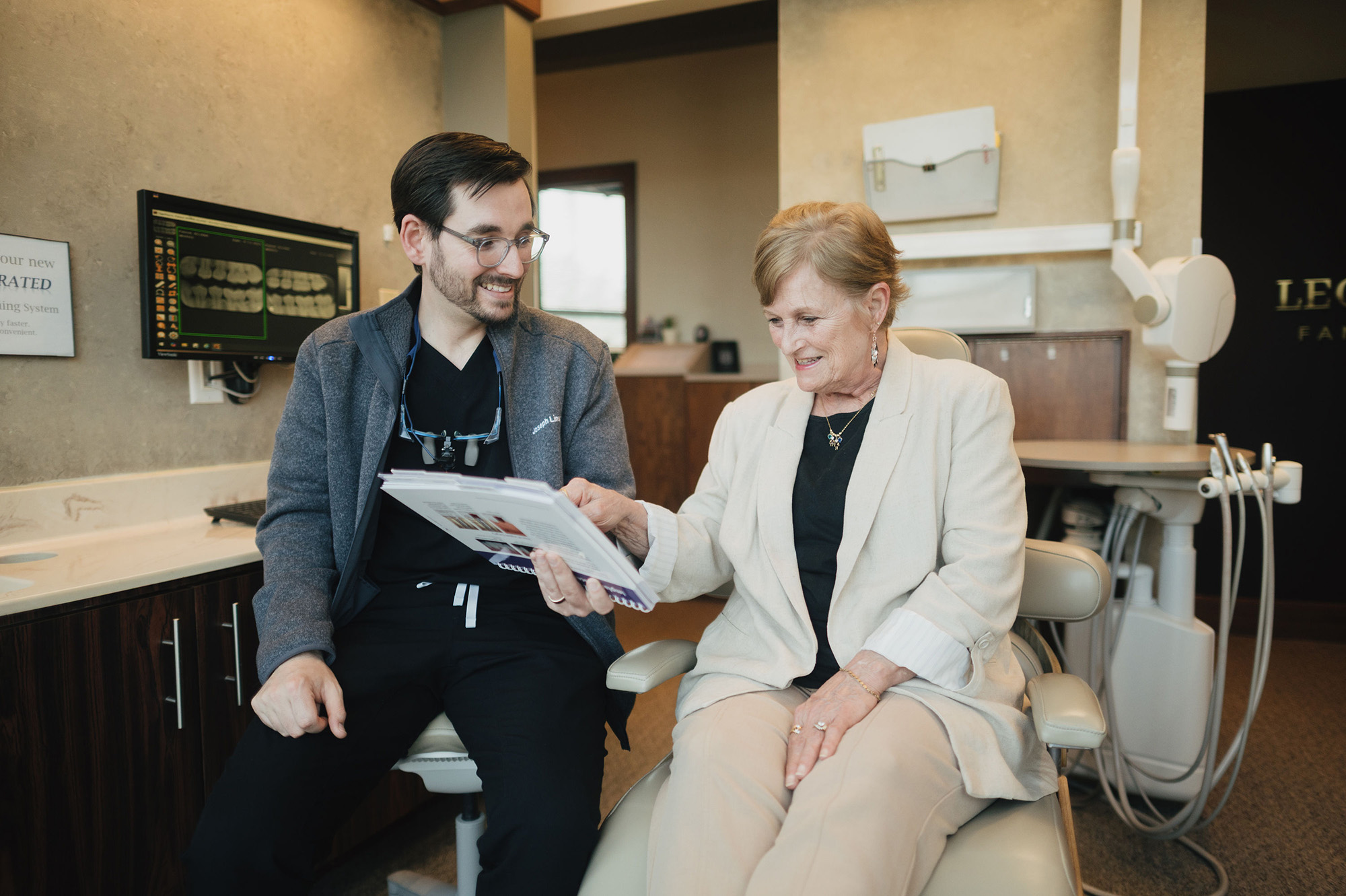 Dentist and older woman review dental documents together in a dental office with X-ray on screen.