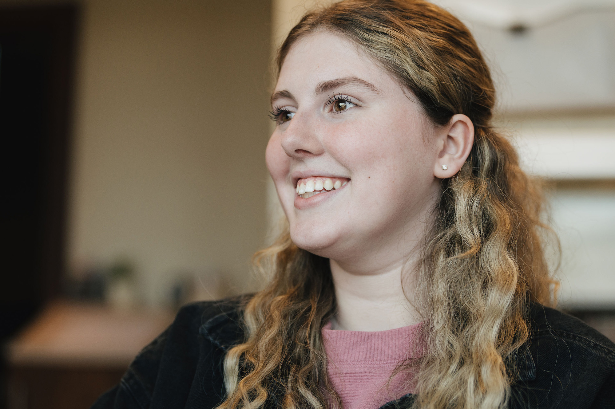 Smiling young woman with wavy blonde hair wearing a pink top and dark jacket indoors.
