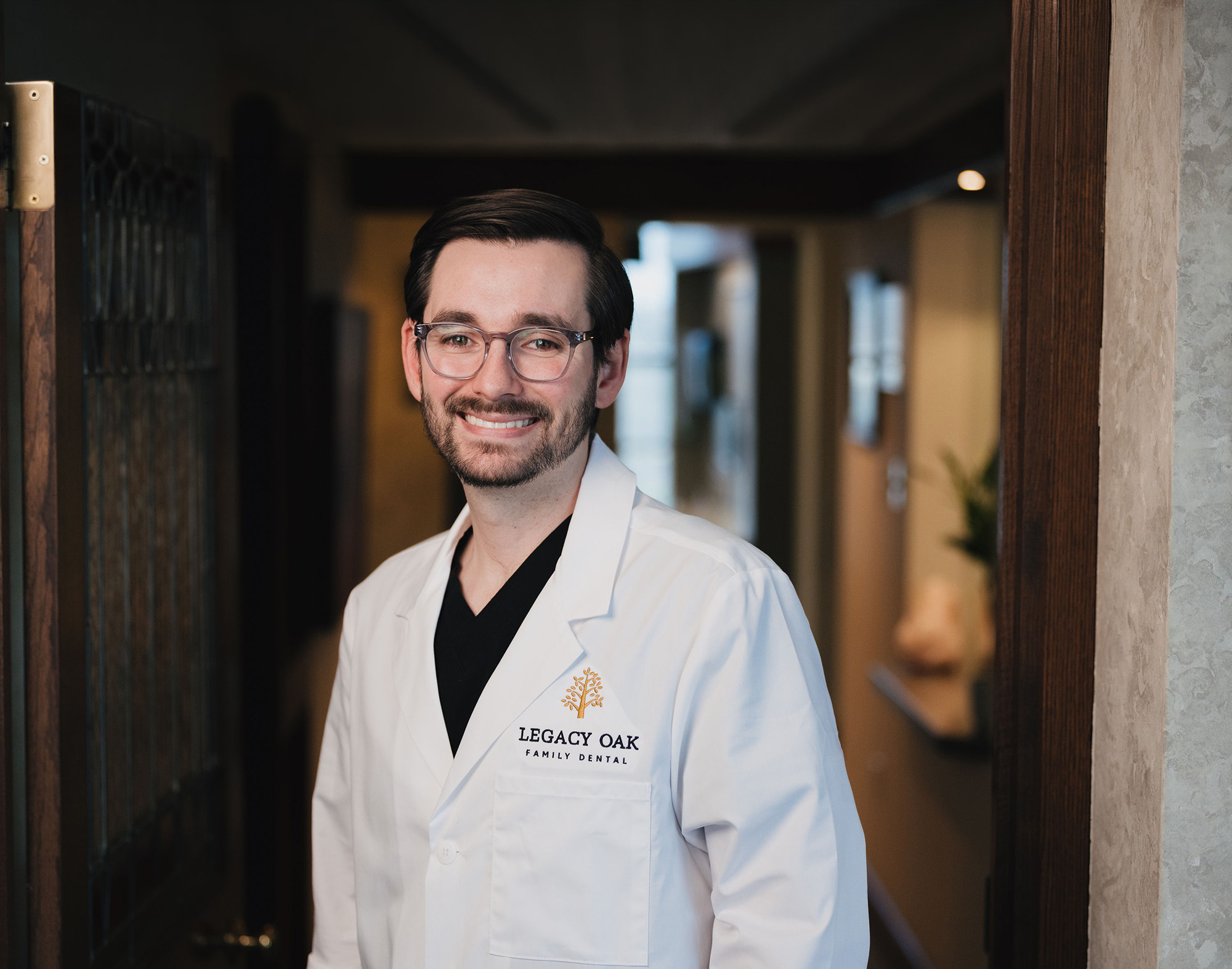 Smiling male dentist wearing glasses and white coat with Legacy Oak Family Dental logo.