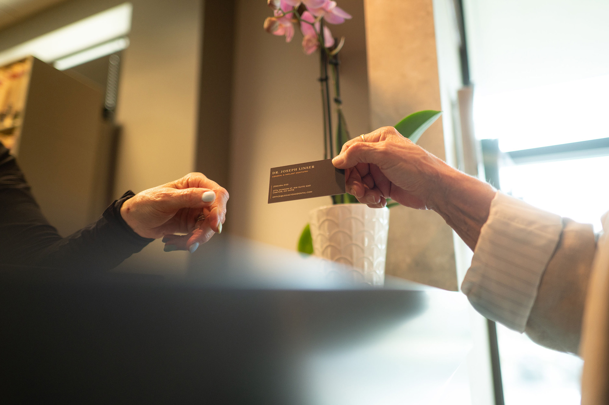 Two people exchanging a business card indoors near a potted orchid with natural light.