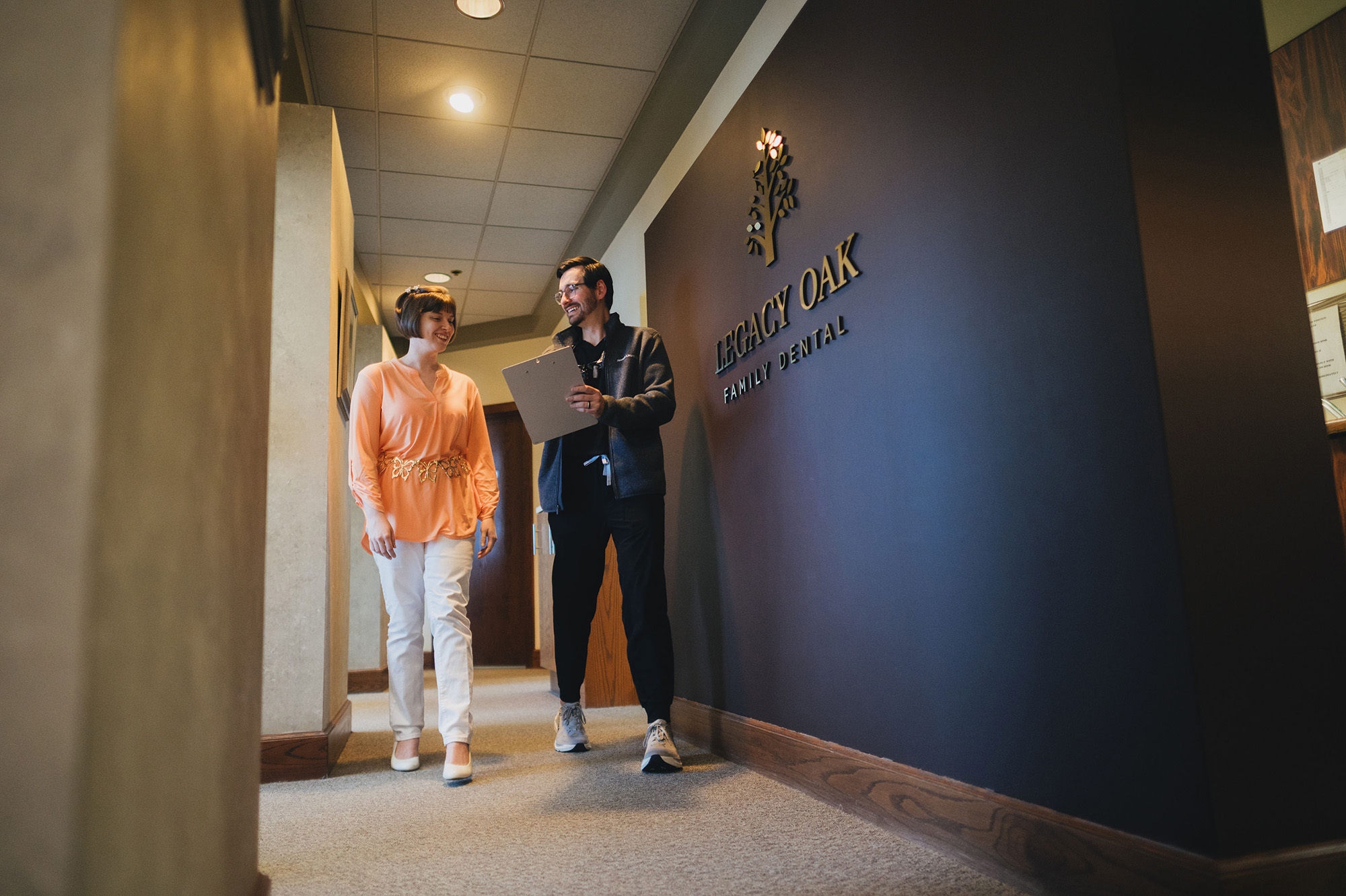 Smiling dental professional walks with female patient along hallway at Legacy Oak Family Dental.