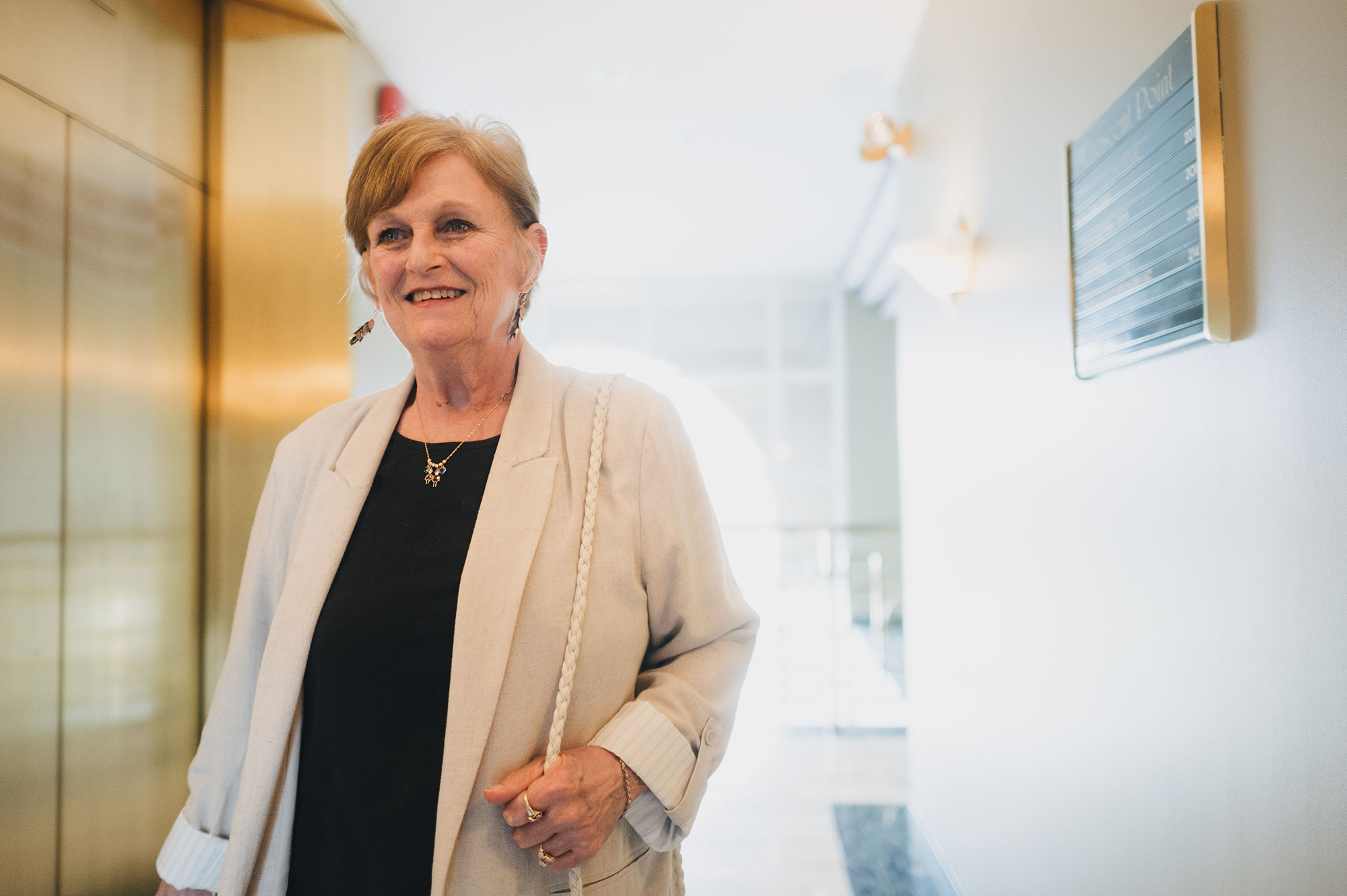 Smiling older woman wearing a light blazer and black top standing in a bright hallway.