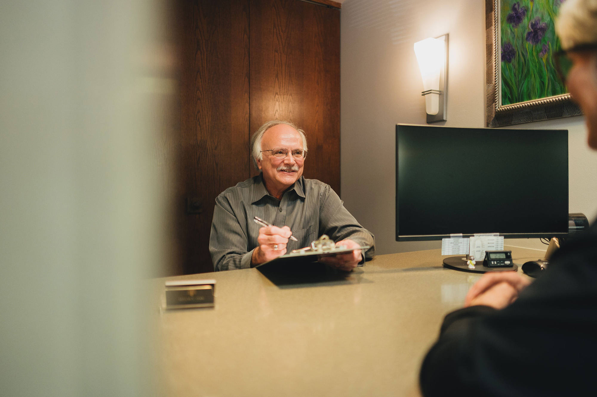 Smiling older man with glasses holding a clipboard, talking to a person at an office desk.