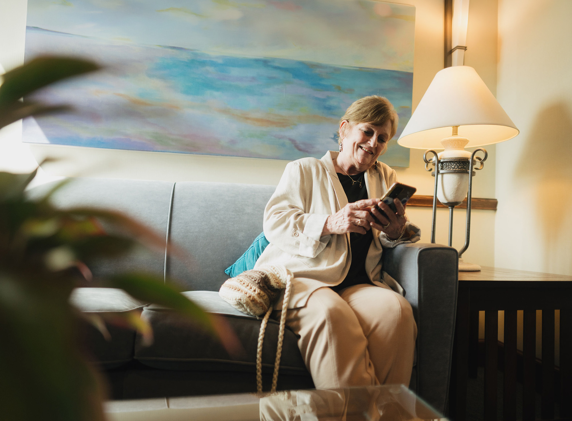 Smiling elderly woman sitting on gray sofa indoors, using a smartphone with a lamp and painting behind her.