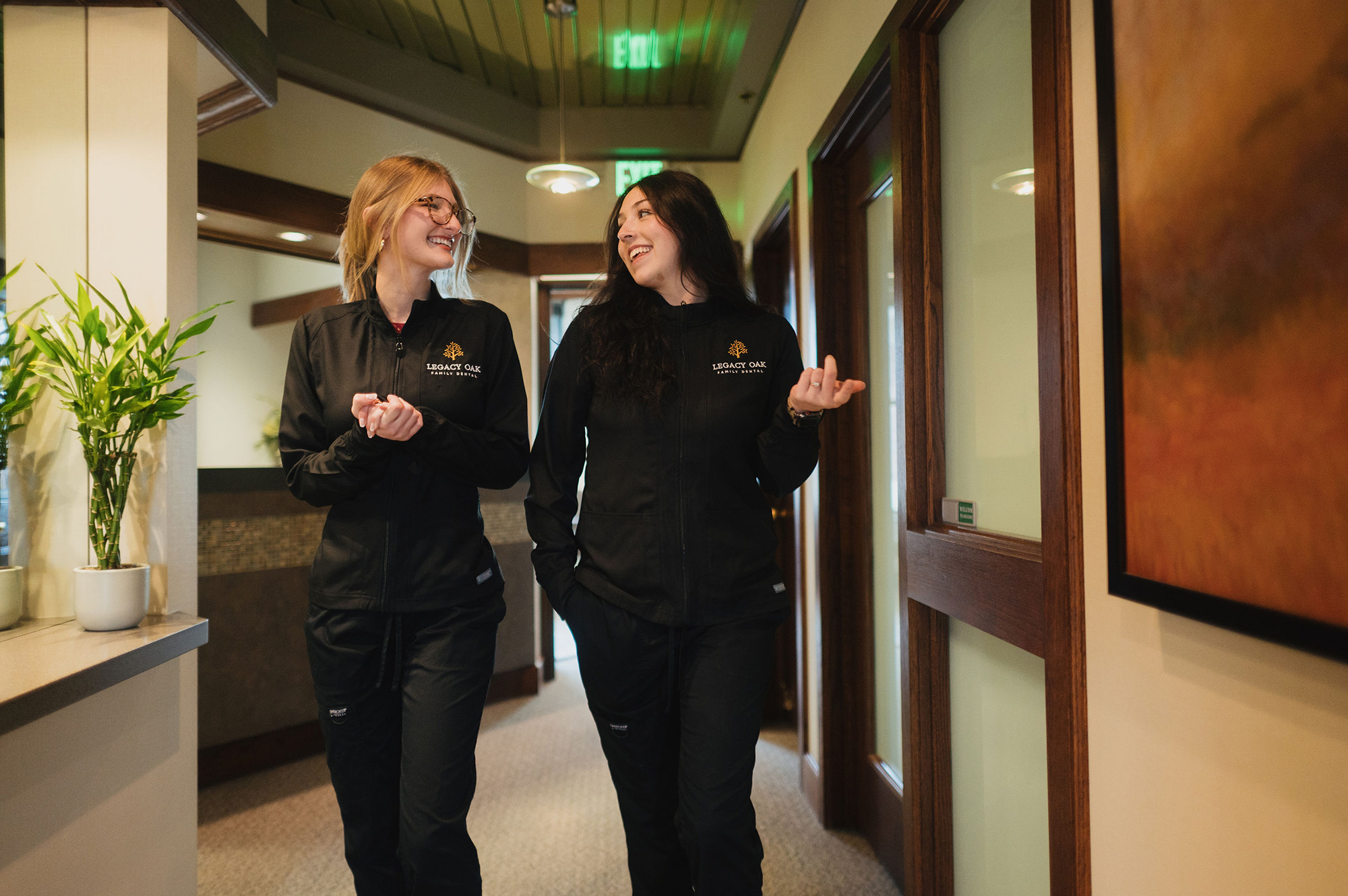 Two women in Legacy Oak Family Dental uniforms walking and smiling in a hallway at work.