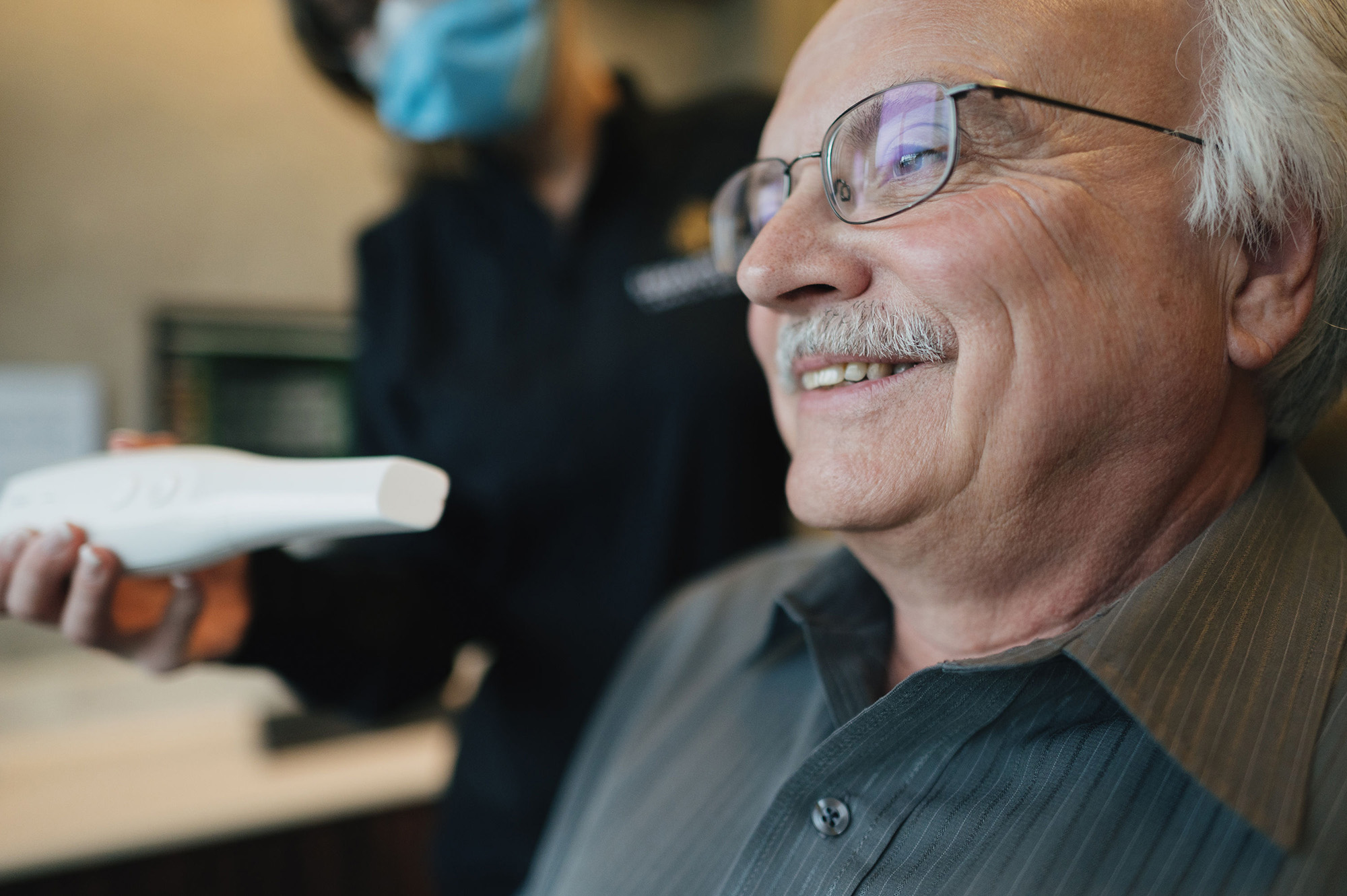 Smiling elderly man wearing glasses, getting a dental scan with a handheld device by a masked professional.