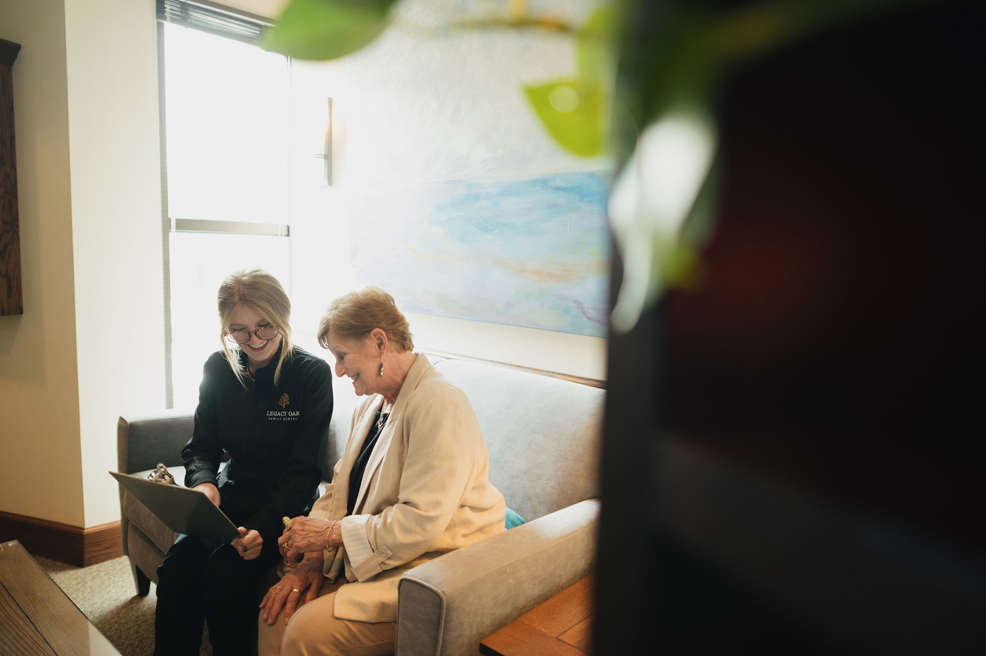 Dental professional and elderly woman smiling while discussing paperwork on a couch in an office.