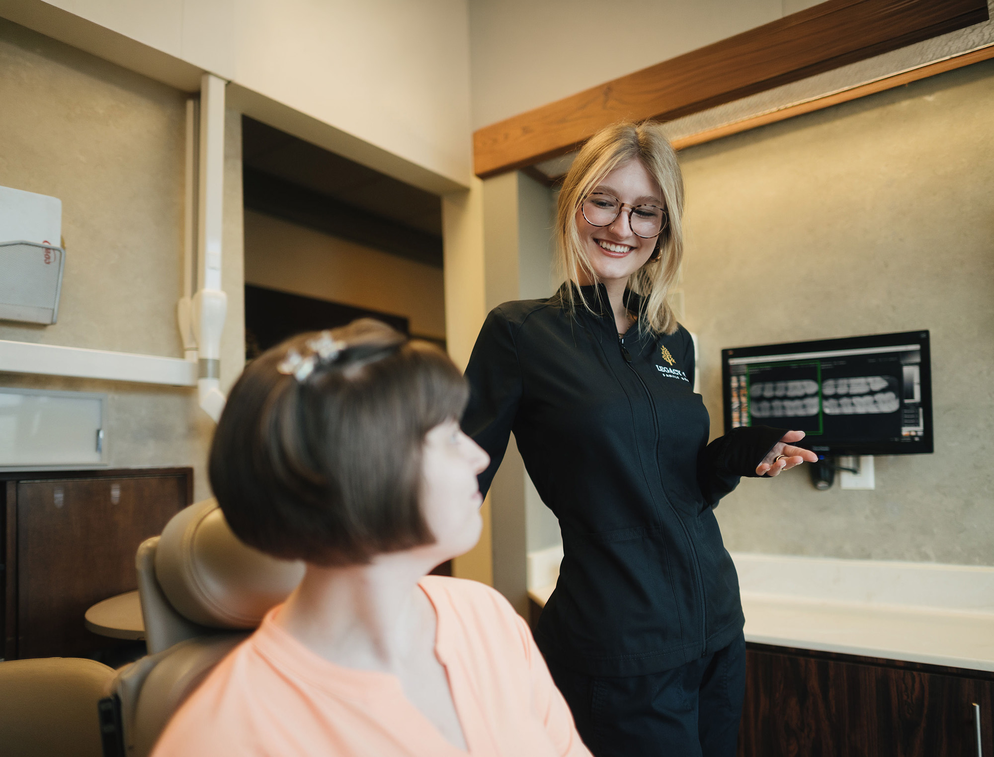 Smiling female dentist in black uniform talks to seated patient with dental X-rays on screen behind.