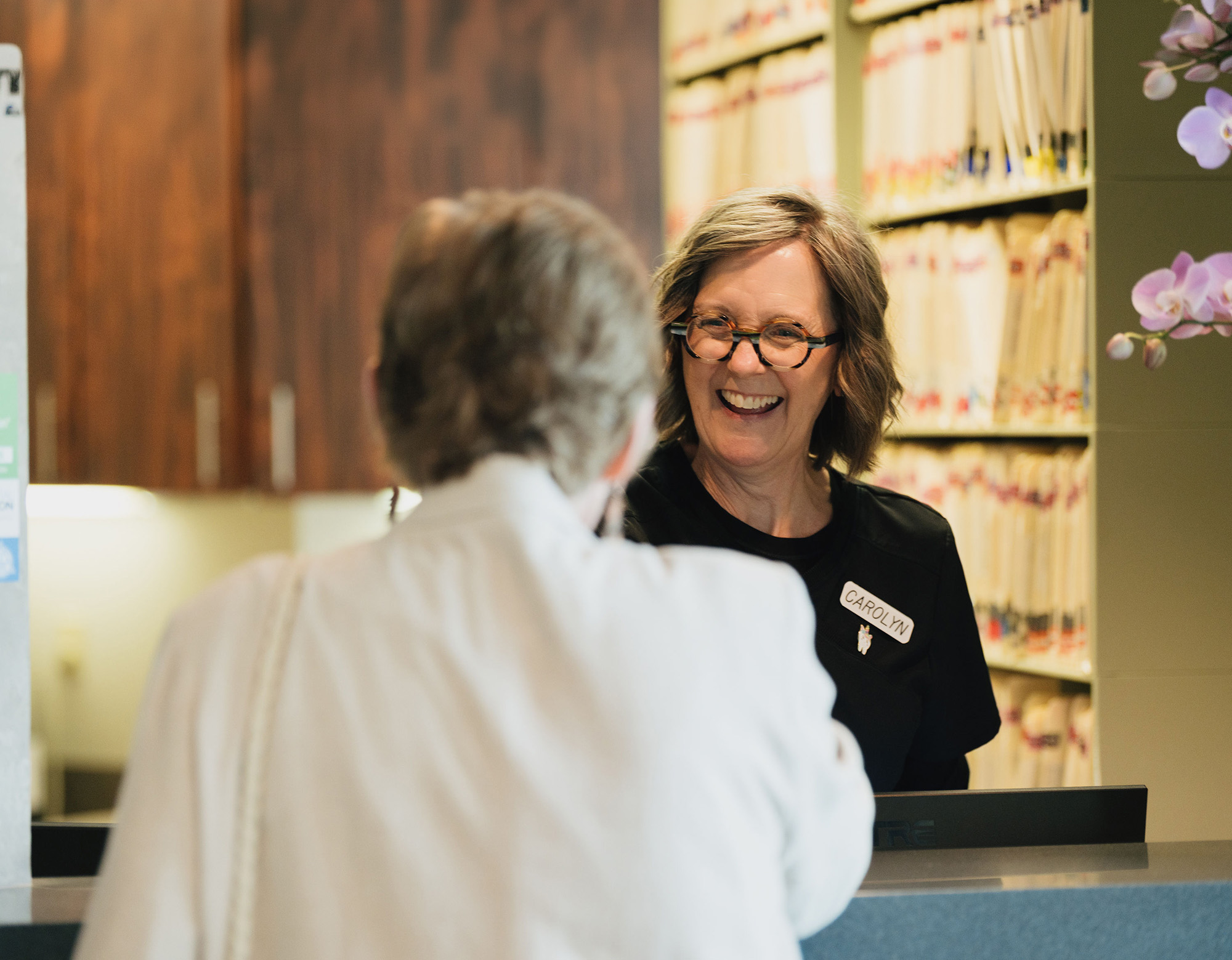 Smiling receptionist named Carolyn greets a patient at the office front desk with files behind her.