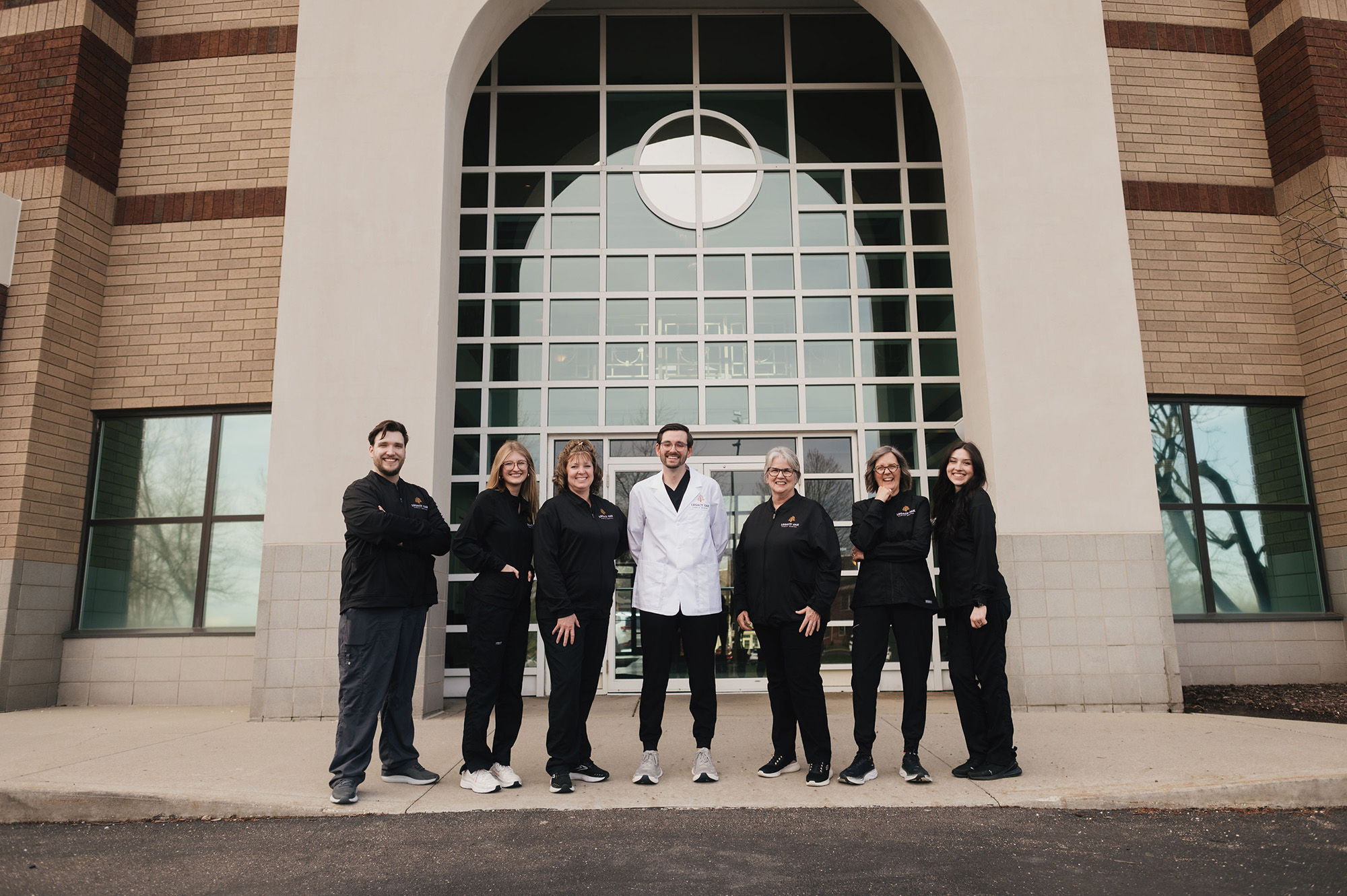 Seven smiling people standing outside a brick building with large windows, one wears a white coat.