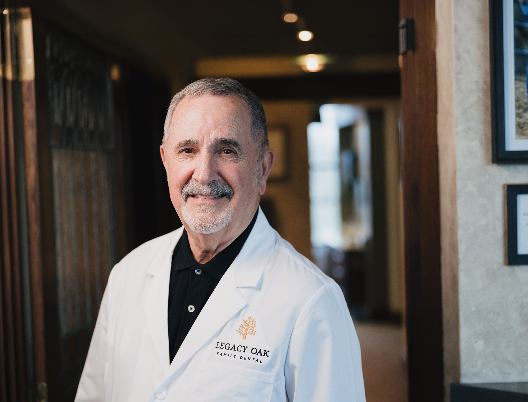 Smiling older man in a white Legacy Oak Family Dental coat standing indoors near a doorway.