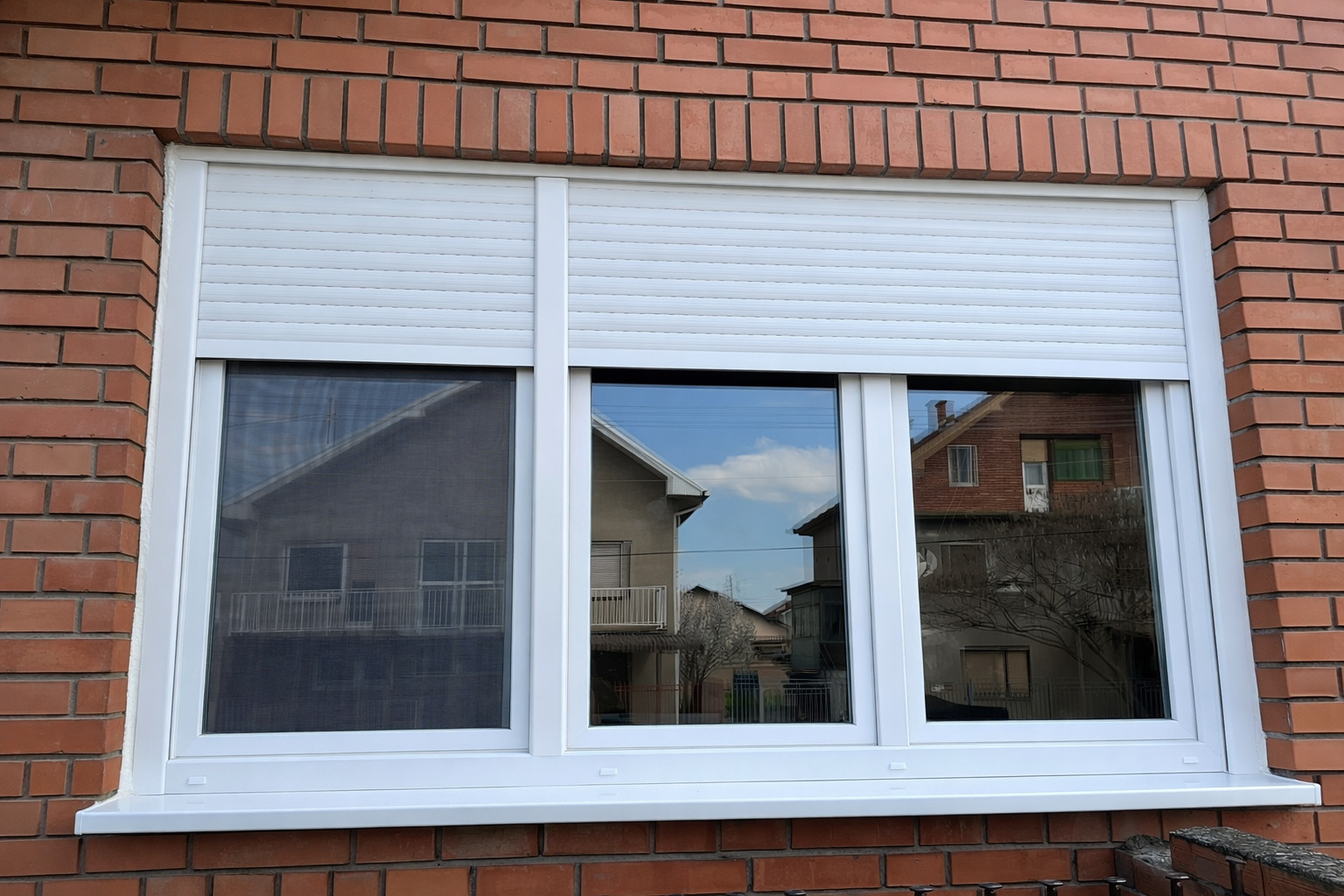 Beli PVC prozor sa spuštenom spoljnom roletnom, ugrađen na fasadi od dekorativne cigle.