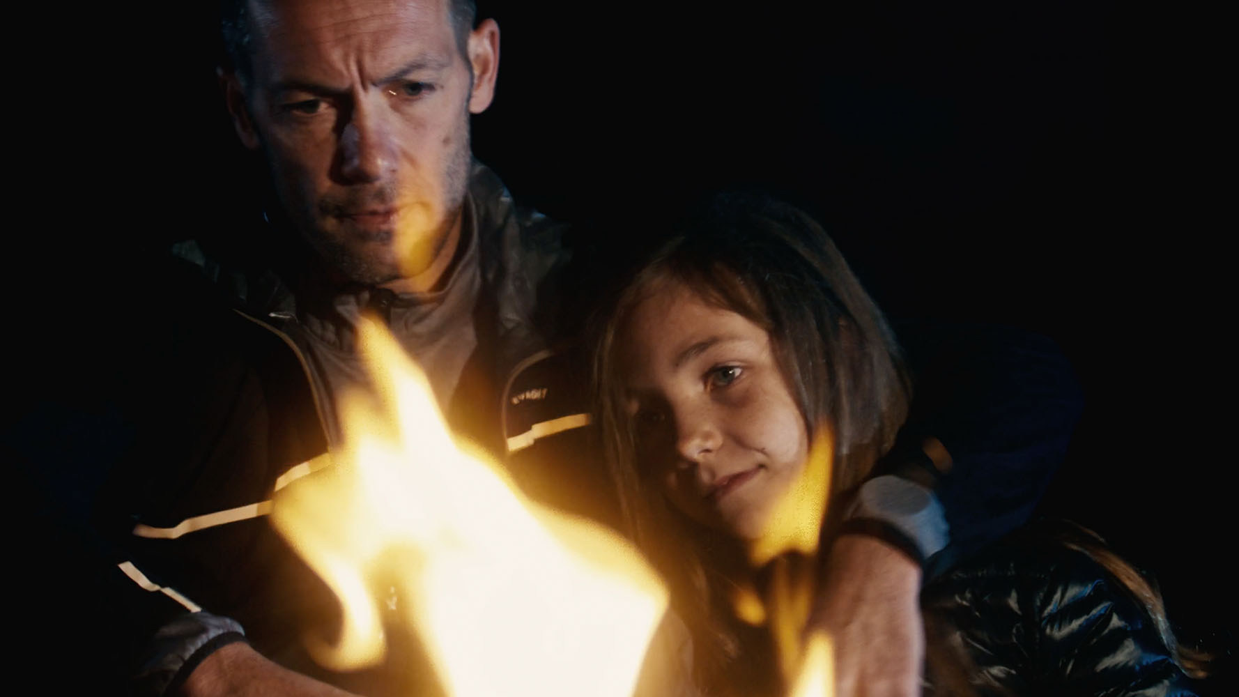 Man and young girl warmly embracing near a campfire at night.