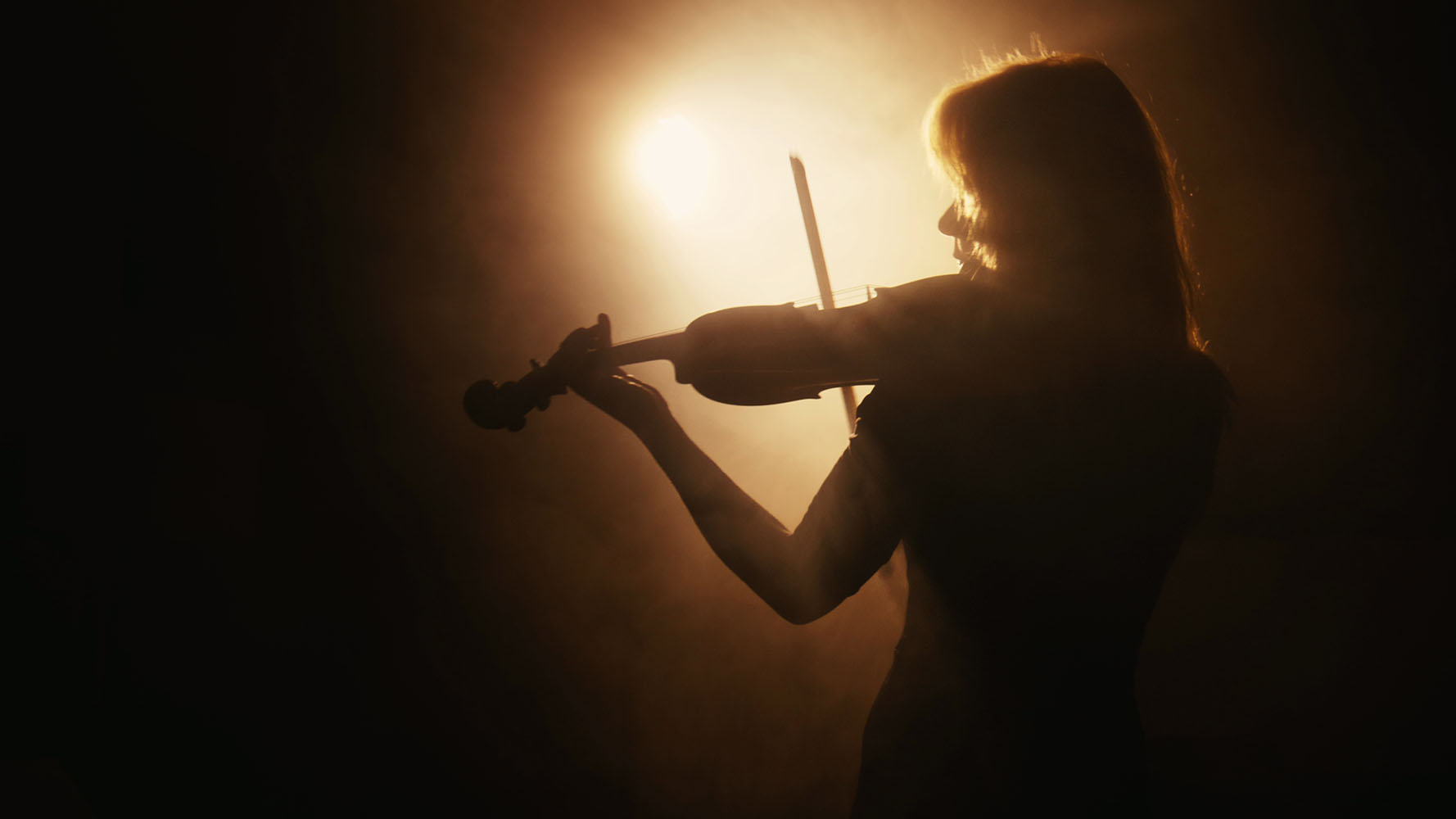 Silhouette of a woman playing violin against a bright backlight in smoky atmosphere.