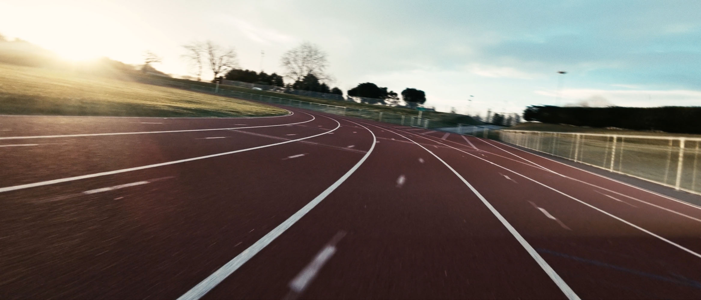 Empty curved outdoor running track under a bright sky with trees and grass surrounding it.