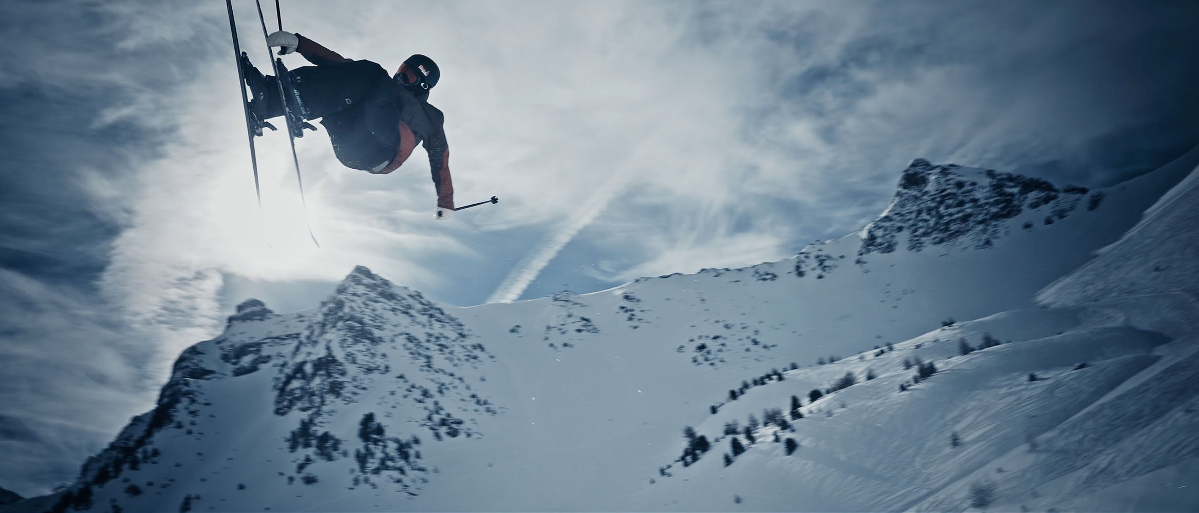 Skier airborne performing a trick in front of snowy mountain peaks under a cloudy sky.