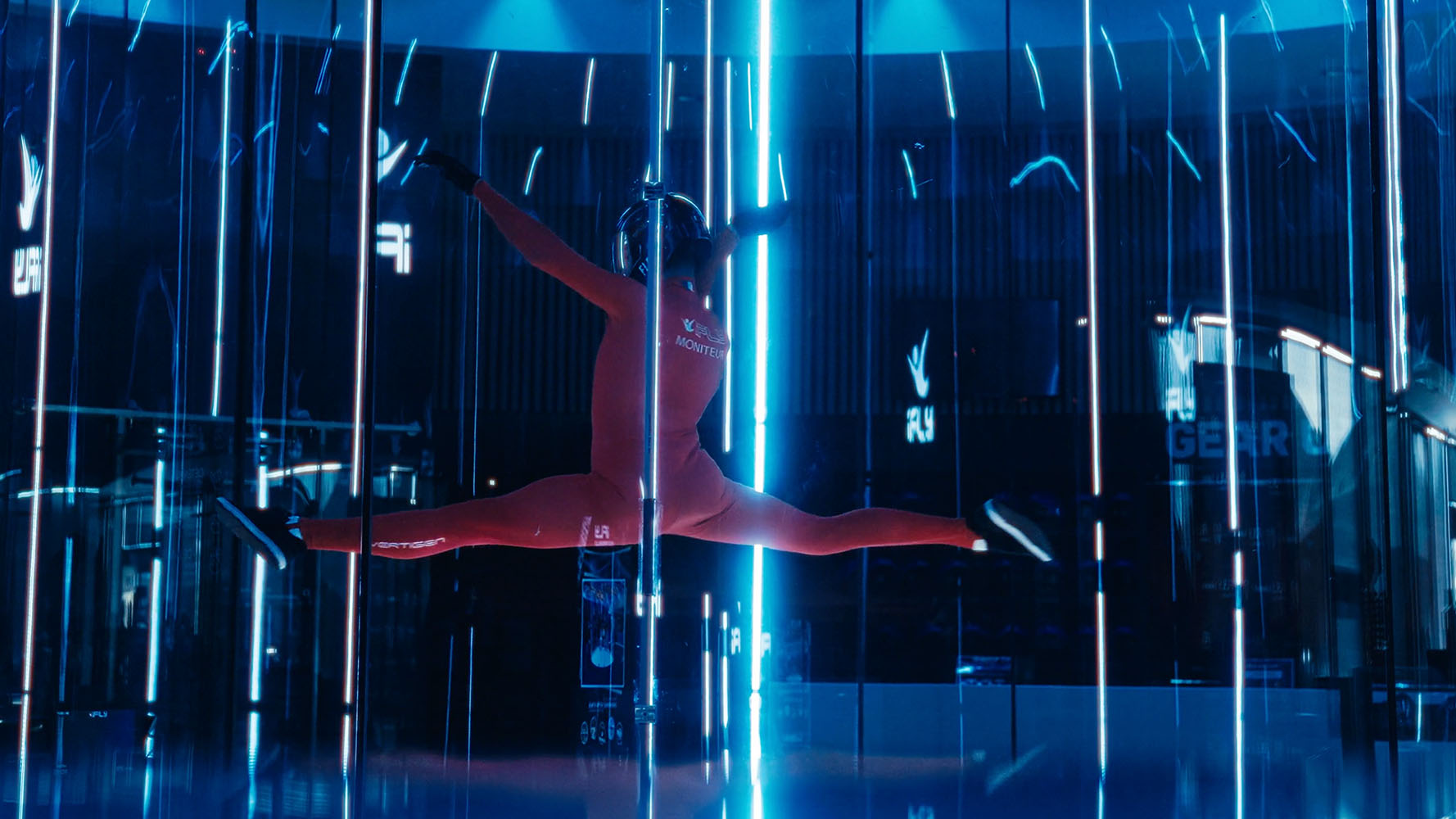 Person in red jumpsuit performing a split jump inside a vertical wind tunnel with blue lighting.