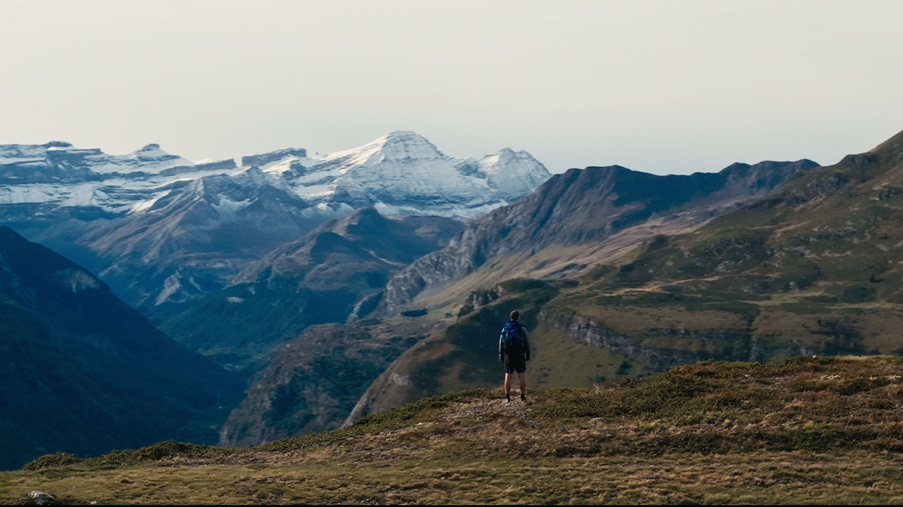 Hiker standing on grassy hill overlooking a mountain range with snow-capped peaks under a clear sky.