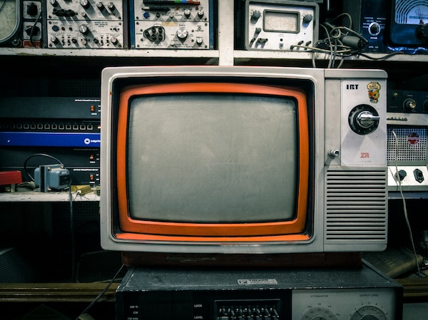 Vintage CRT television with orange border and control knobs on right side, surrounded by electronic equipment.