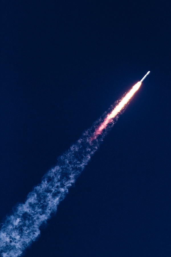 Rocket launching with bright flames and smoke trail against dark blue sky.
