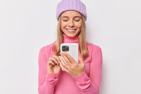 Smiling young woman wearing a pink turtleneck and lavender knit hat using a smartphone.