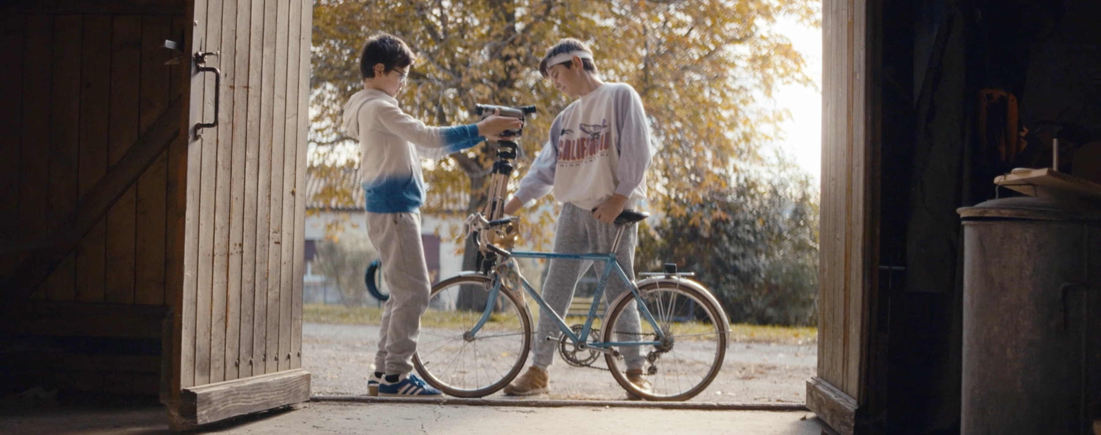 Two boys working on a bicycle inside a wooden shed with autumn trees outside.