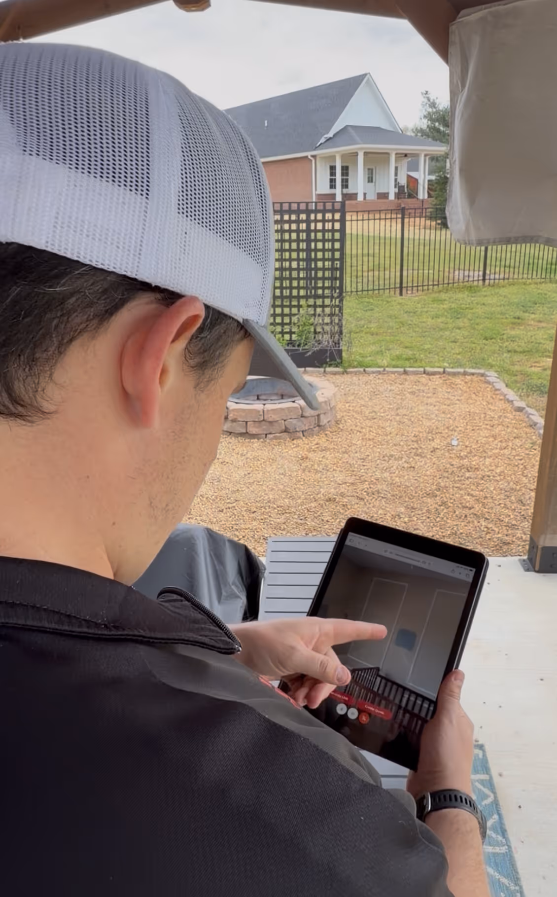 Man wearing a white mesh cap pointing at a tablet screen while standing on a patio with a backyard and house in the background.