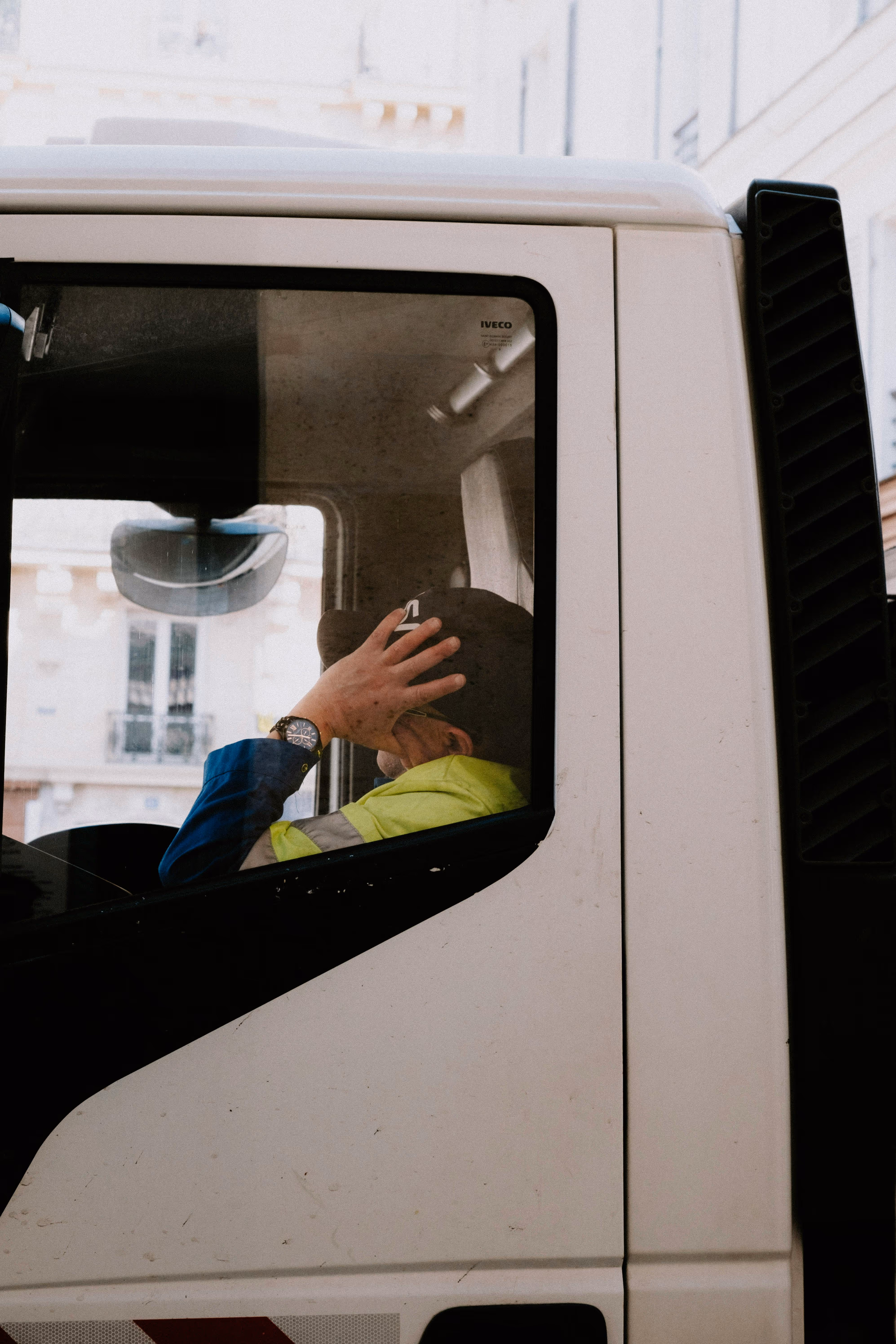A man wearing a high-visibility jacket and cap is sitting inside a white Iveco truck, resting his hand on his head.