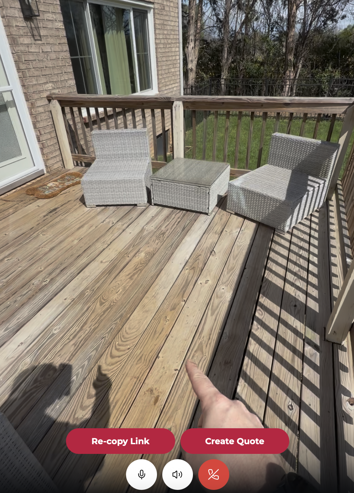 Wooden deck with two gray wicker chairs and a matching square table, with a person's hand pointing towards the deck boards.