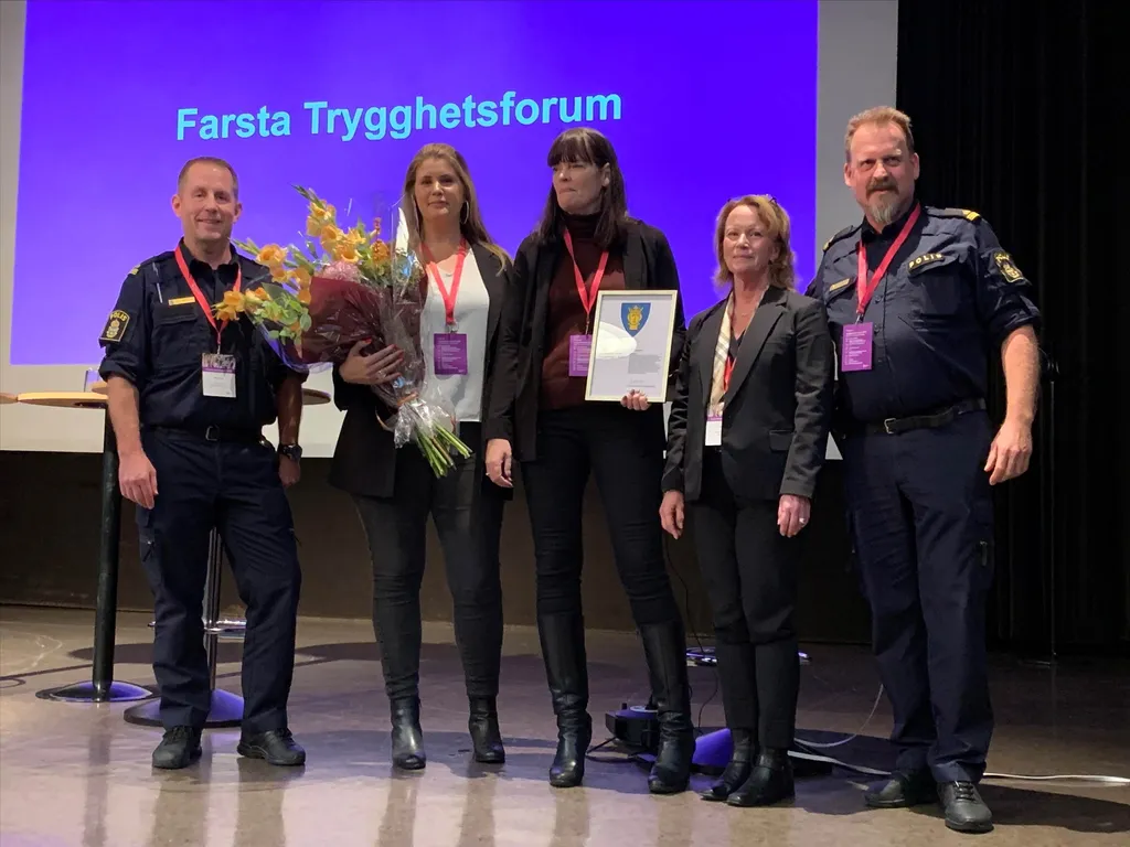 Fem personer pa scen framfor skarmen Farsta Trygghetsforum, tva i polisuniform, en med blommor och en med diplom.
