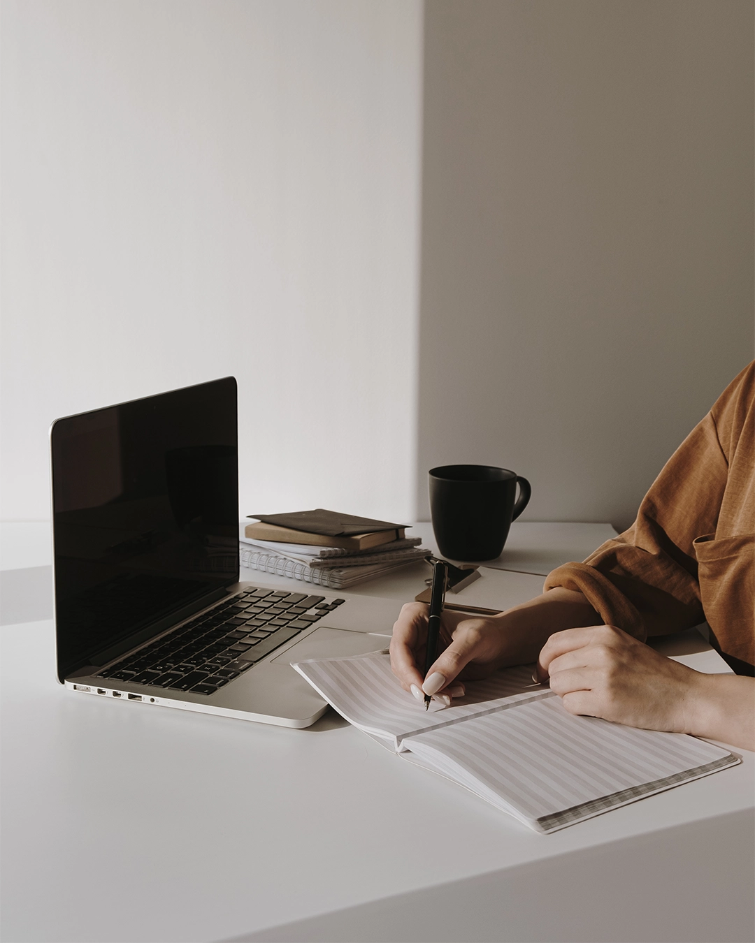 Person schreibt mit einem Stift in ein offenes Notizbuch neben einem Laptop, Stapel Bücher und eine schwarze Tasse auf einem weißen Schreibtisch.