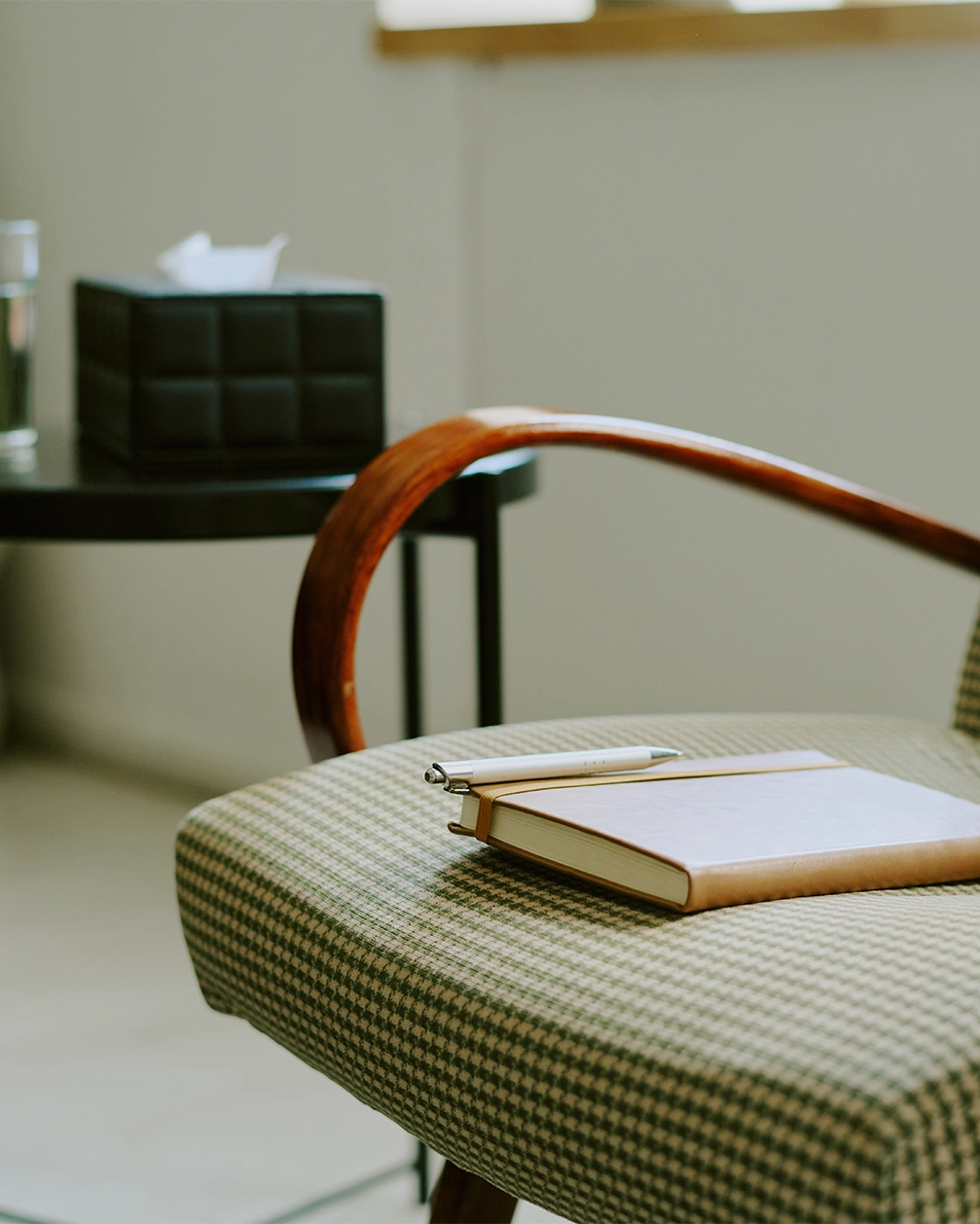 Sessel mit kariertem Stoffbezug, auf dem ein Notizbuch und ein Stift liegen, daneben ein Tisch mit einer Box Papiertücher und einem Glas Wasser.
