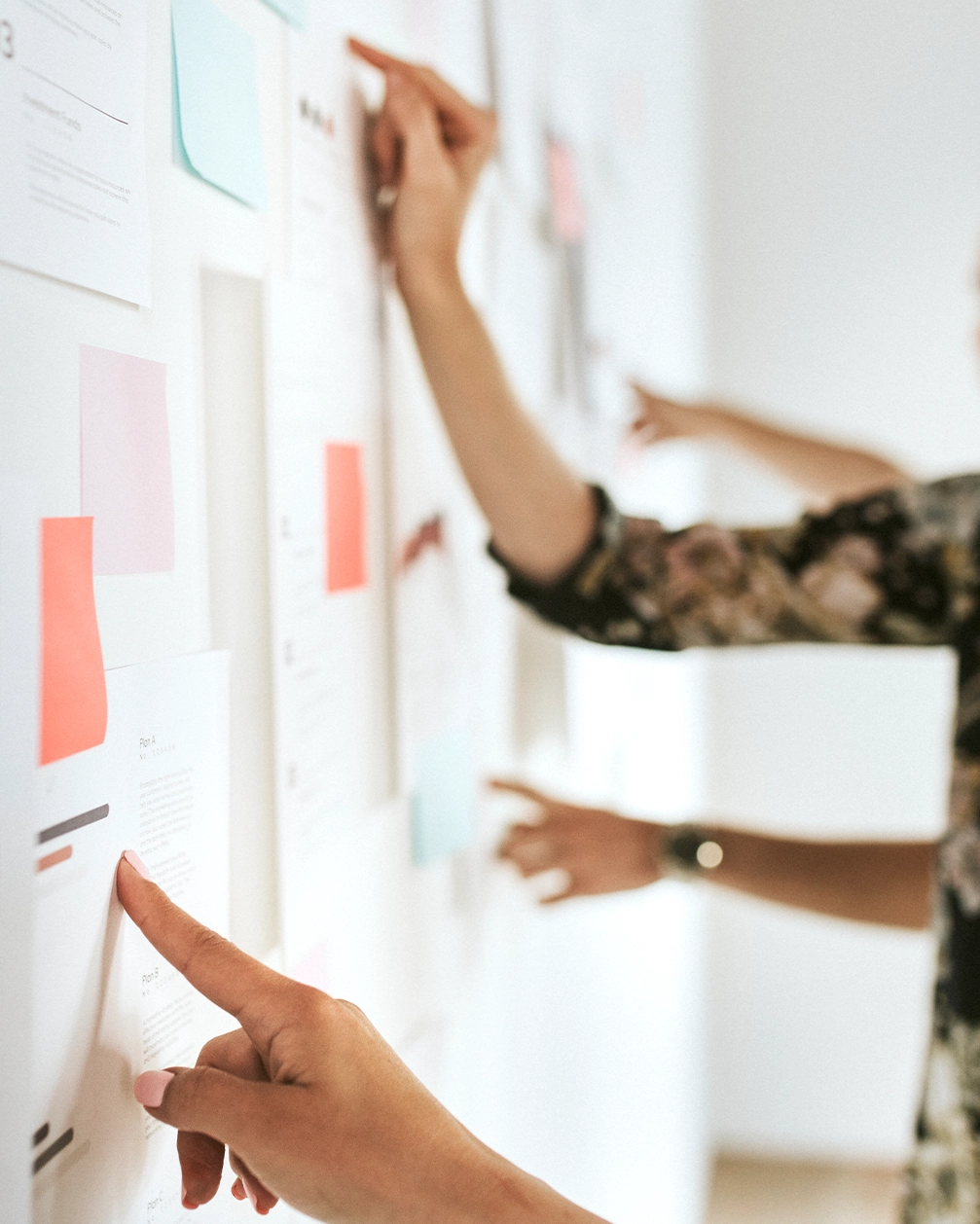 Person zeigt mit Finger auf verschiedene auf einer Wand angeheftete bunte Post-it-Notizen und Papiere in einem Besprechungsraum.