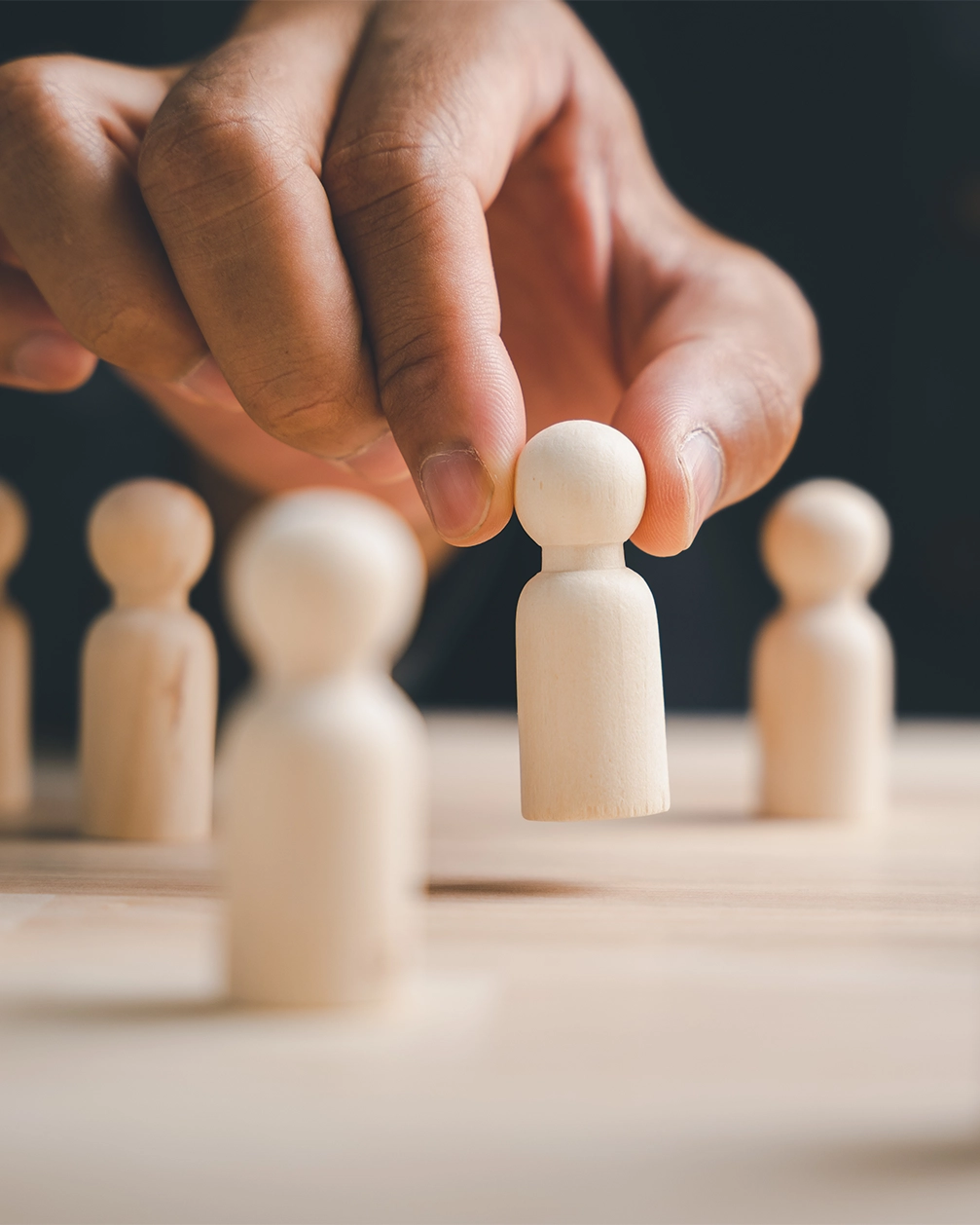 Hand, die eine hölzerne Spielfigur von einem Tisch aufhebt, mit mehreren ähnlichen Figuren im Hintergrund.