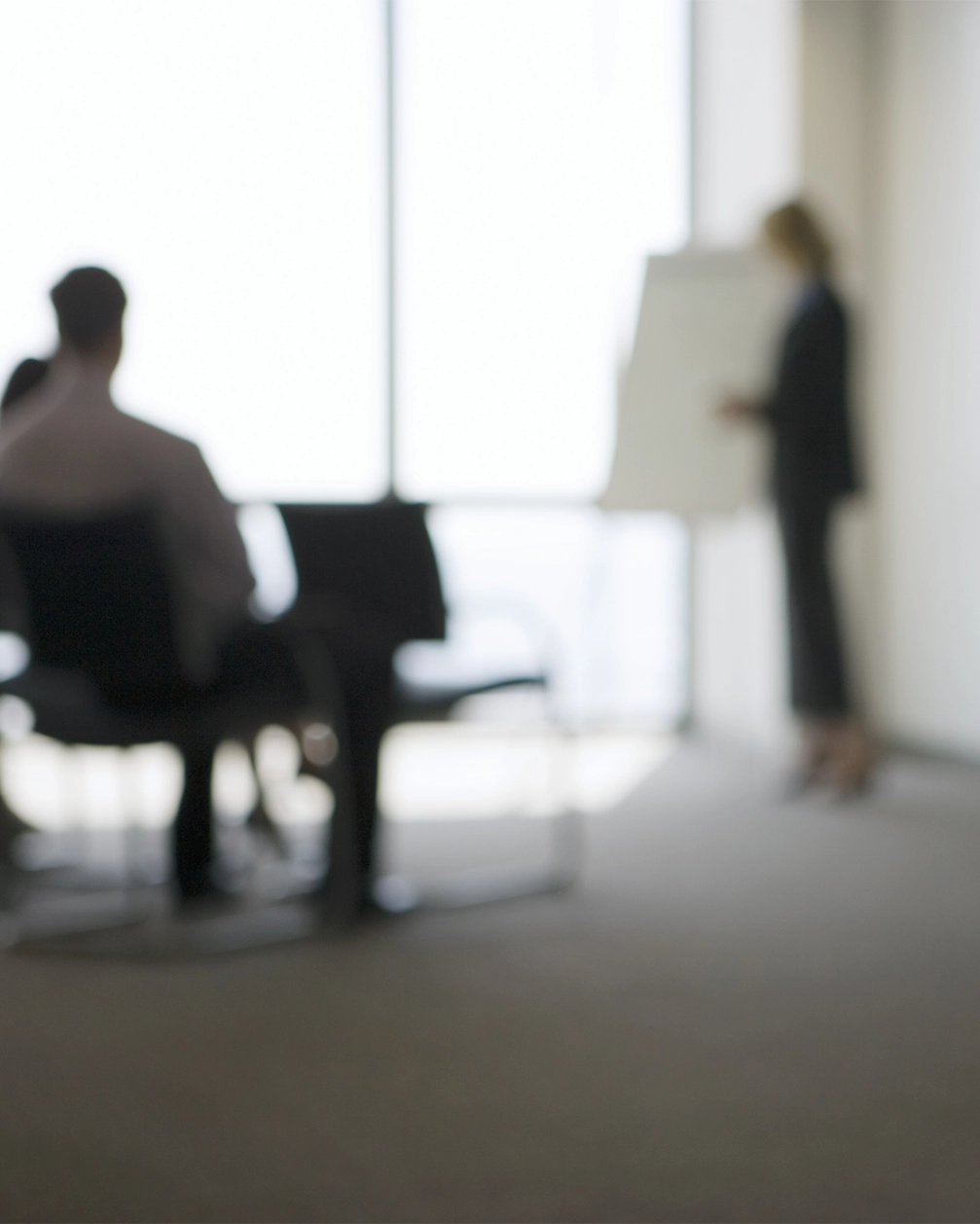 Verschwommenes Bild einer Frau, die vor einem Whiteboard in einem Büro spricht, während Zuhörer auf Stühlen sitzen.
