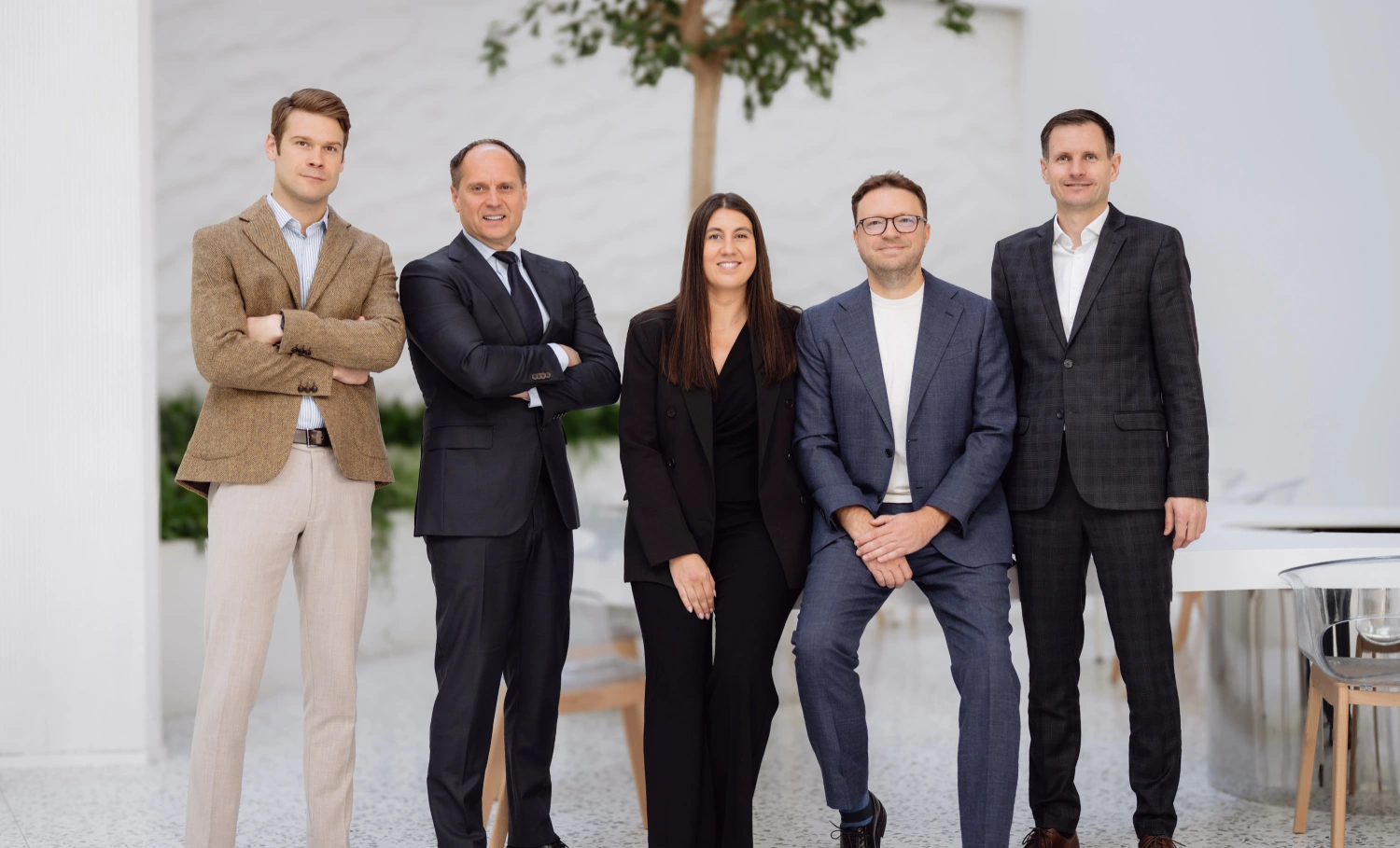 Five professionally dressed people standing indoors, posing for a group photo with neutral background and some plants.