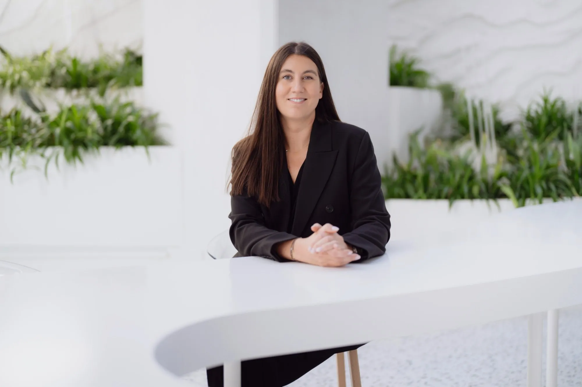 A woman with long dark hair wearing a black blazer sits at a white curved table with green plants in the background.