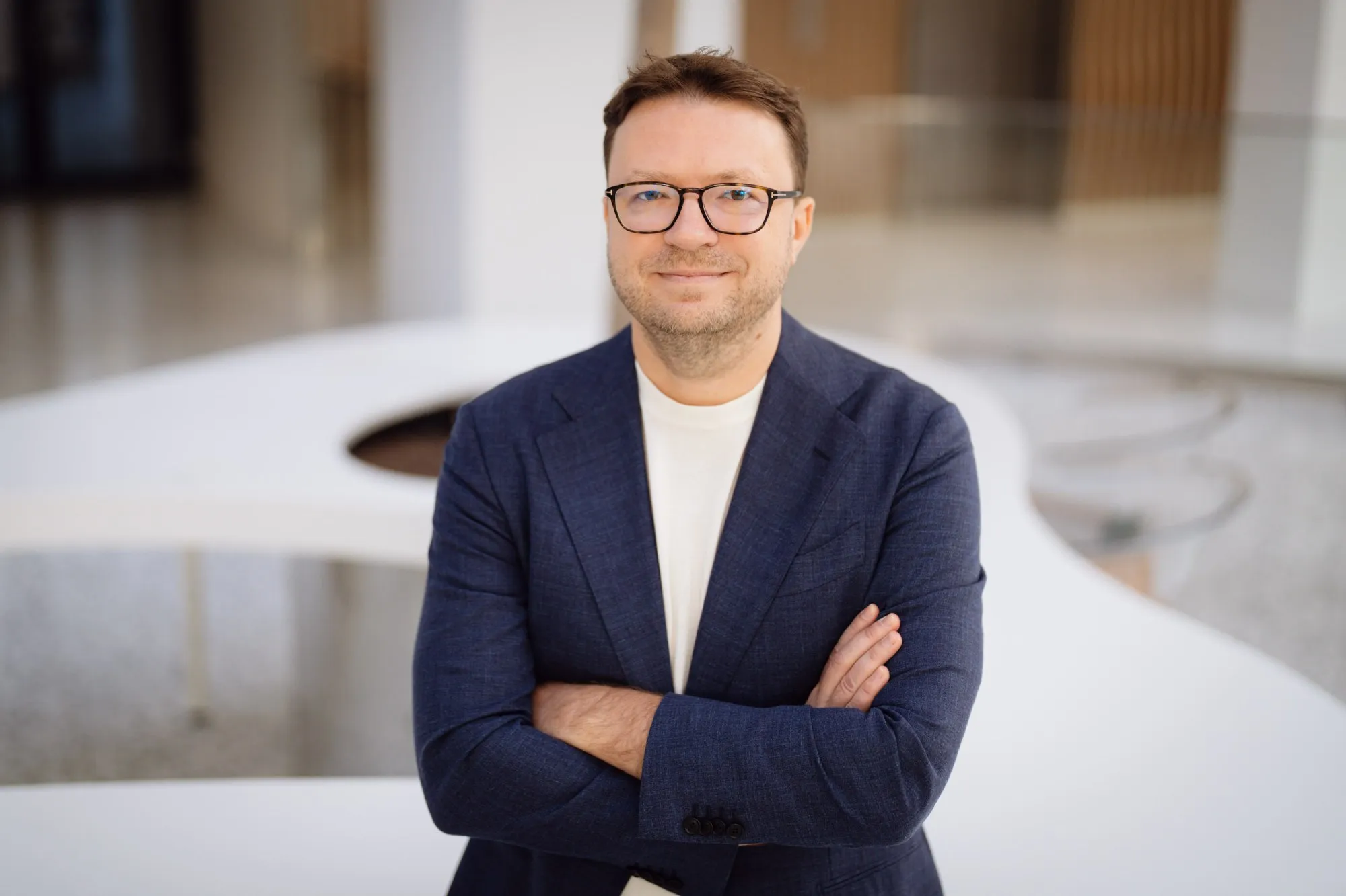 Man with glasses wearing a navy blazer and white shirt, standing with arms crossed in a modern, bright indoor setting.