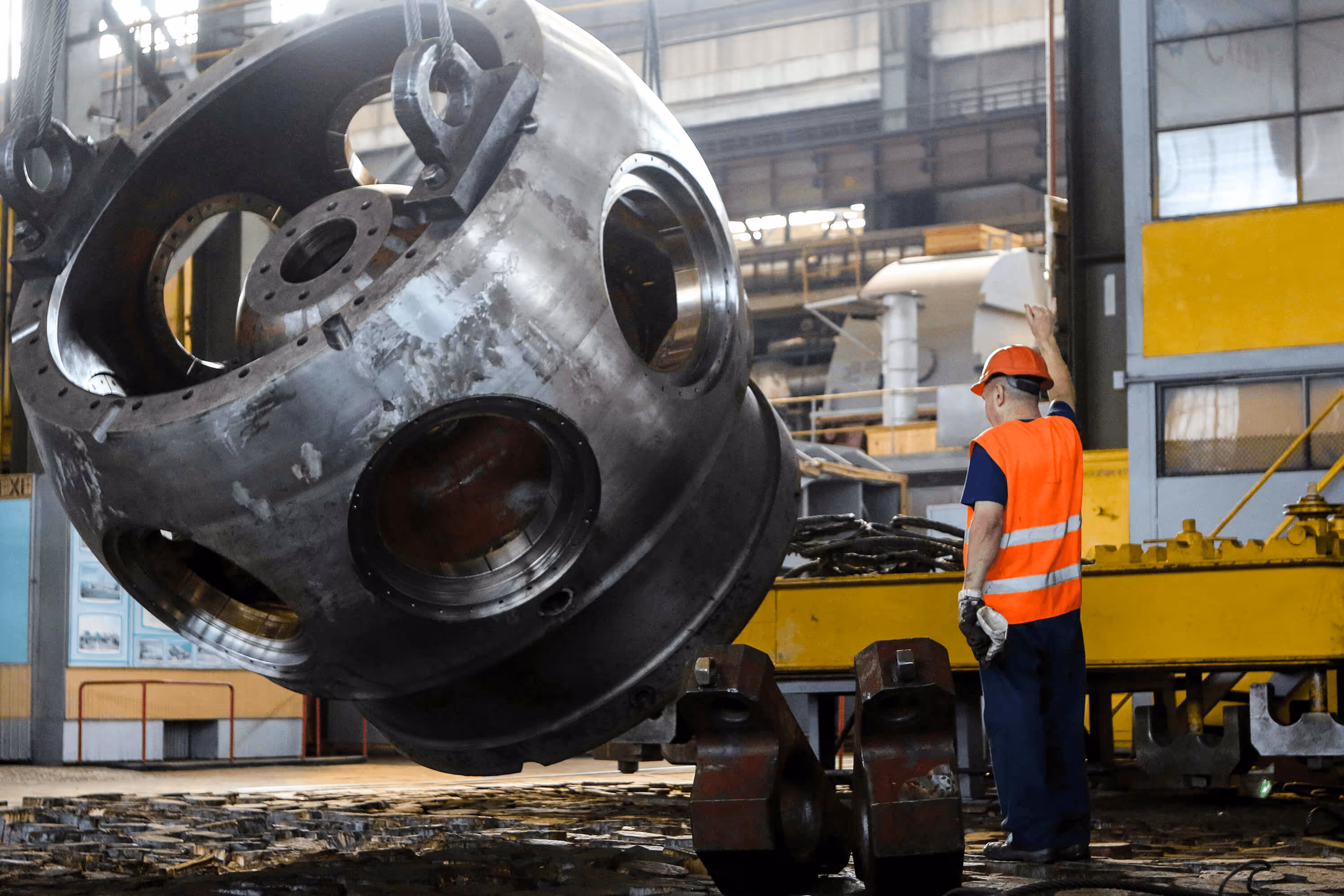 Worker in orange safety vest and hard hat guiding a large industrial metal component suspended by cables inside a factory.