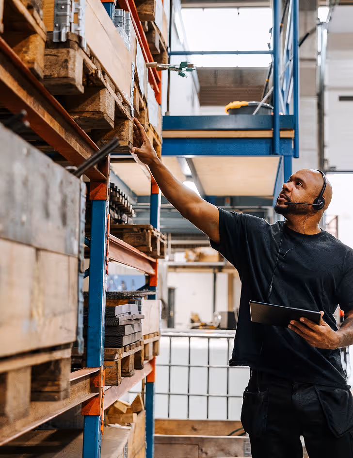 Warehouse worker wearing headset and holding a tablet reaching for items on a high shelf in an industrial storage area.
