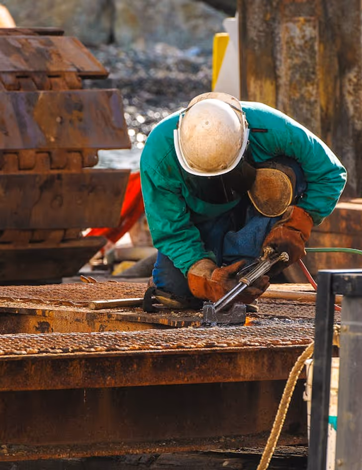 Construction worker wearing a white hard hat and green jacket kneeling and using a cutting torch on metal grating.