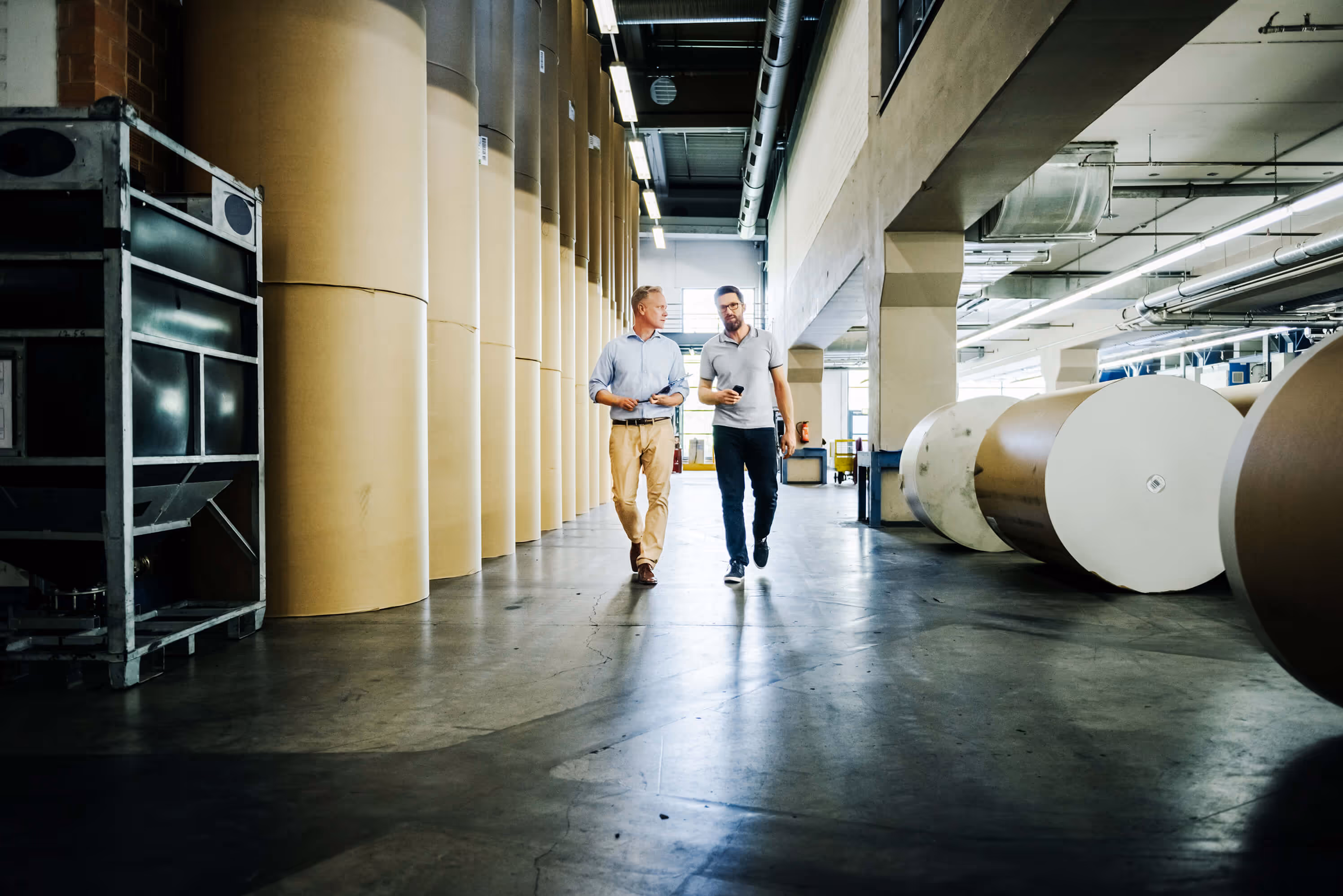 Two men walking and talking inside a large paper manufacturing or storage facility with large rolls of paper and industrial equipment.